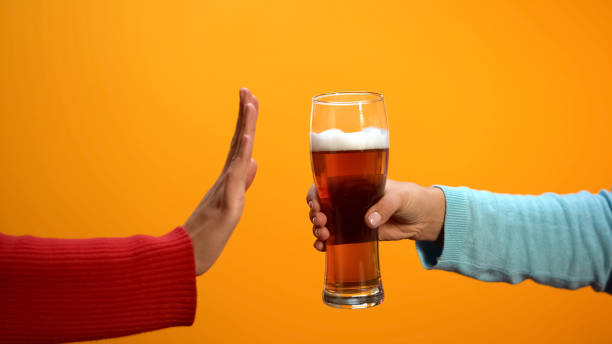 Obraz przedstawia symboliczne odrzucenie napoju alkoholowego, sugerując odmowę lub decyzję o zaprzestaniu spożywania alkoholu. Użyte kolory – żółty i ciemne – podkreślają kontrast między proponowanym drinkiem a decyzją o jego braku.
