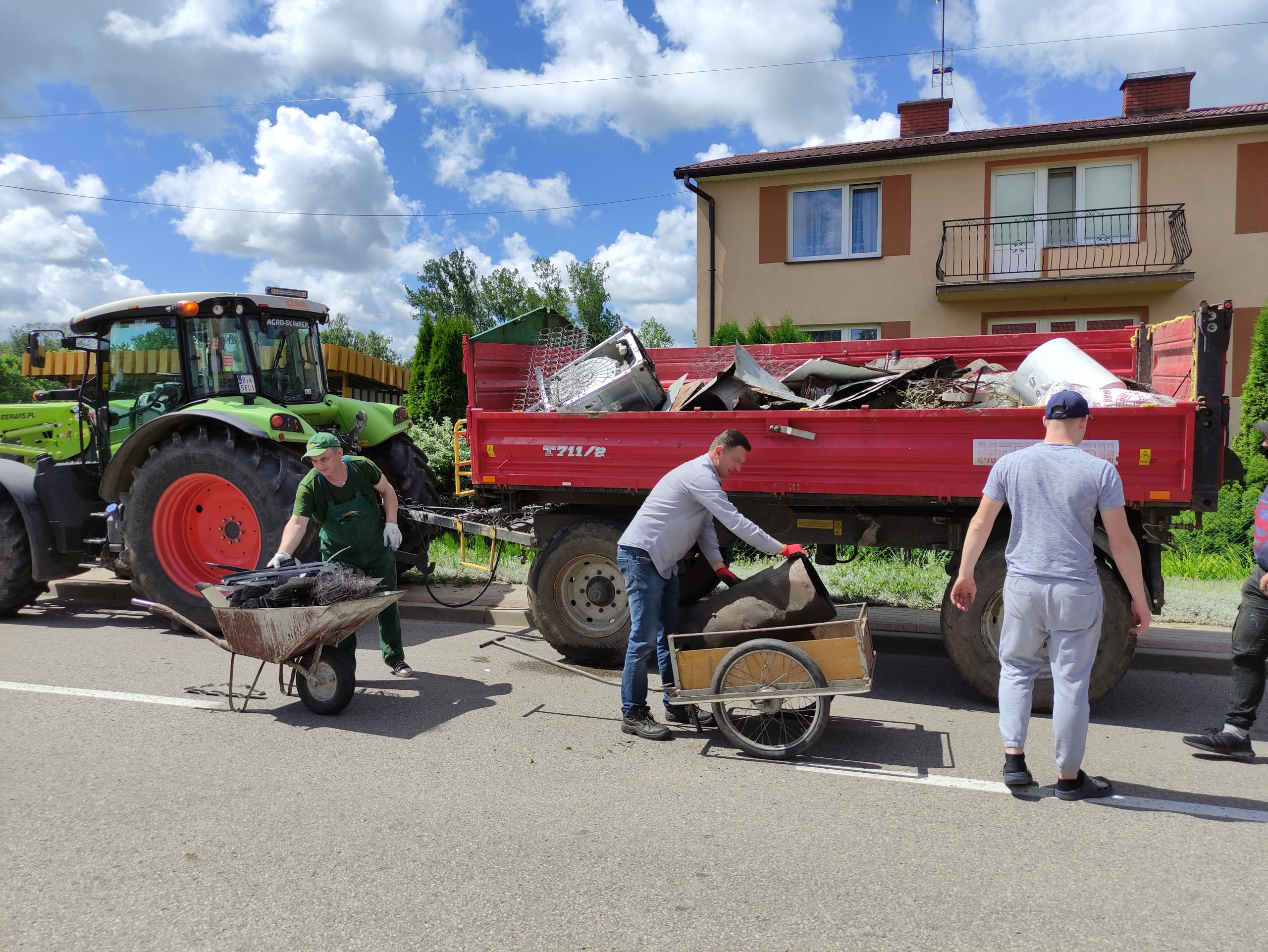 Mężczyźni zbierają złom na potrzeby budowy Domu Miłosierdzia w gminie Wyszki. Na przyczepie ciągnionej przez traktor znajduje się gruz, a obok stoi wiadro na śmieci.”