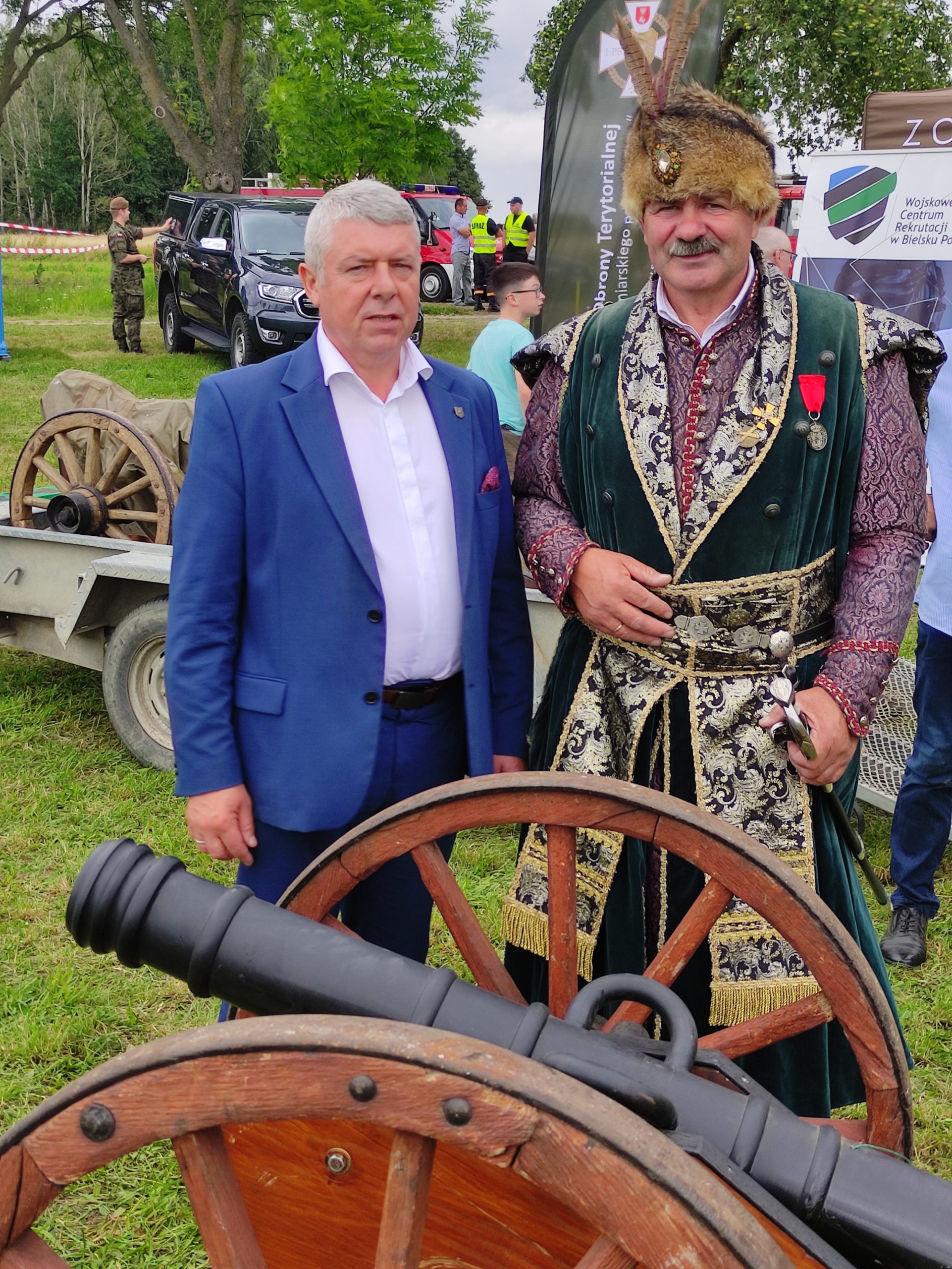 Dwoje osób – elegancki mężczyzna w garniturze i osoba w historycznym stroju – stoi obok działa podczas wydarzenia. W tle widoczni są inni ludzie i pojazdy.