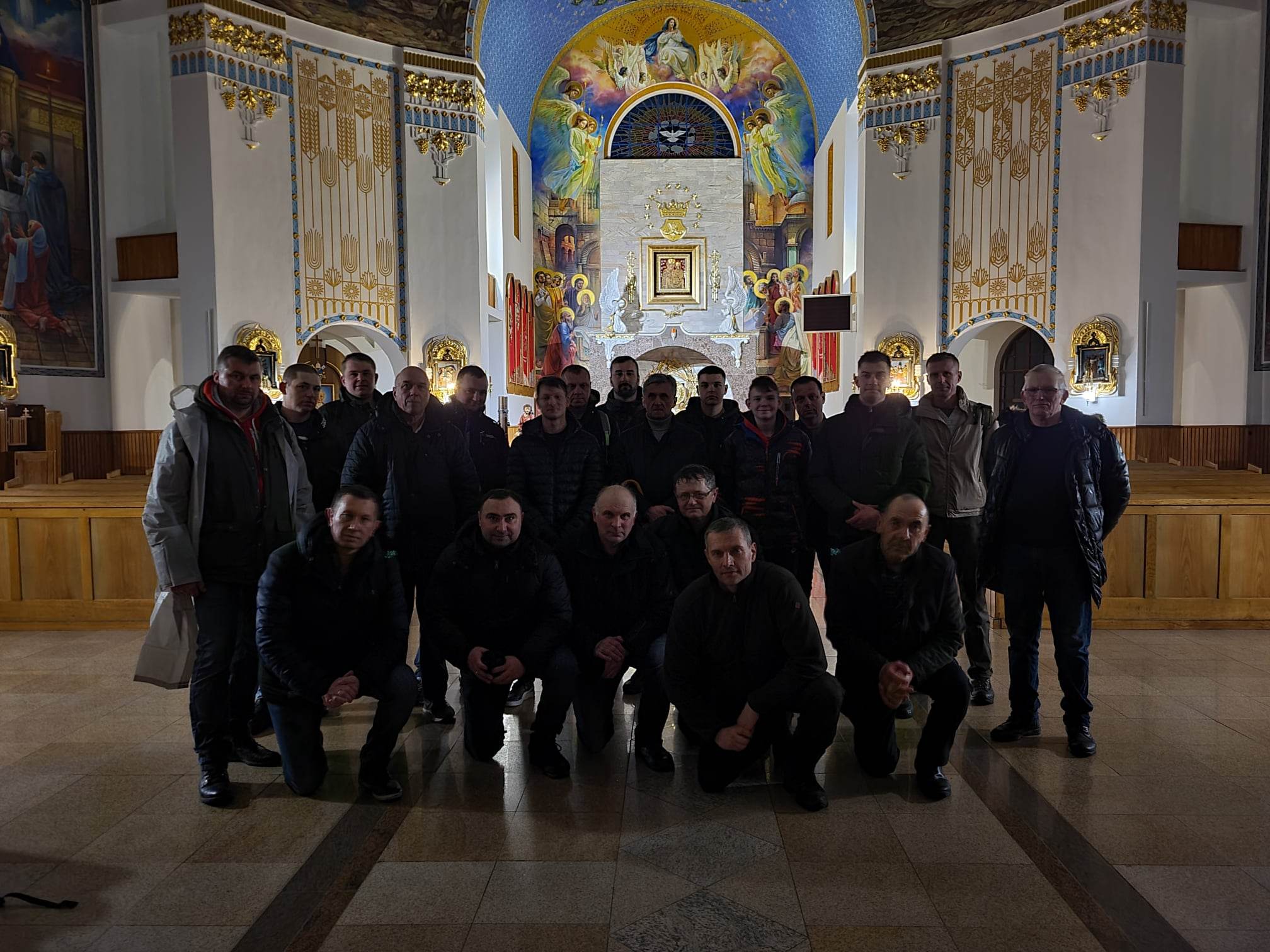 Grupa osób stoi wewnątrz kościoła. W tle znajduje się duża, kolorowa mozaika przedstawiająca postać na krzyżu. Osoby są ubrane w ciemne ubrania.