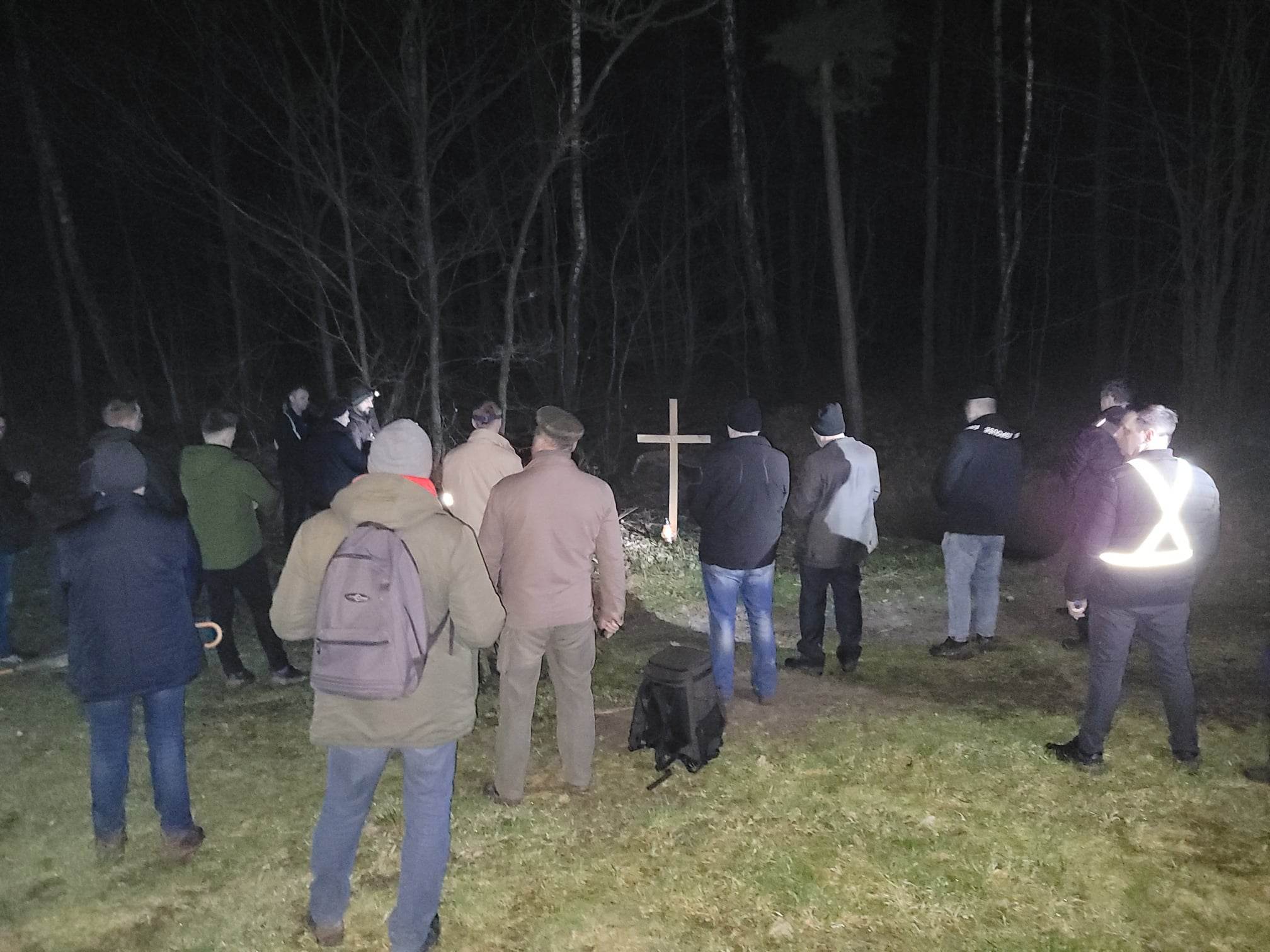 W nocy grupa mężczyzn bierze udział w nocnej drodze krzyżowej, otoczona drzewami i krzyżami. Niektóre osoby mają latarnie lub świece.