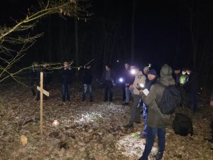 Grupa osób w ciemnym lesie, używa latarek do oświetlenia drewnianego krzyża postawionego na ziemi. Wokół krzyża leżą liście i gałęzie. Osoby są ubrane w różne ubrania, głównie kurtki i czapki.