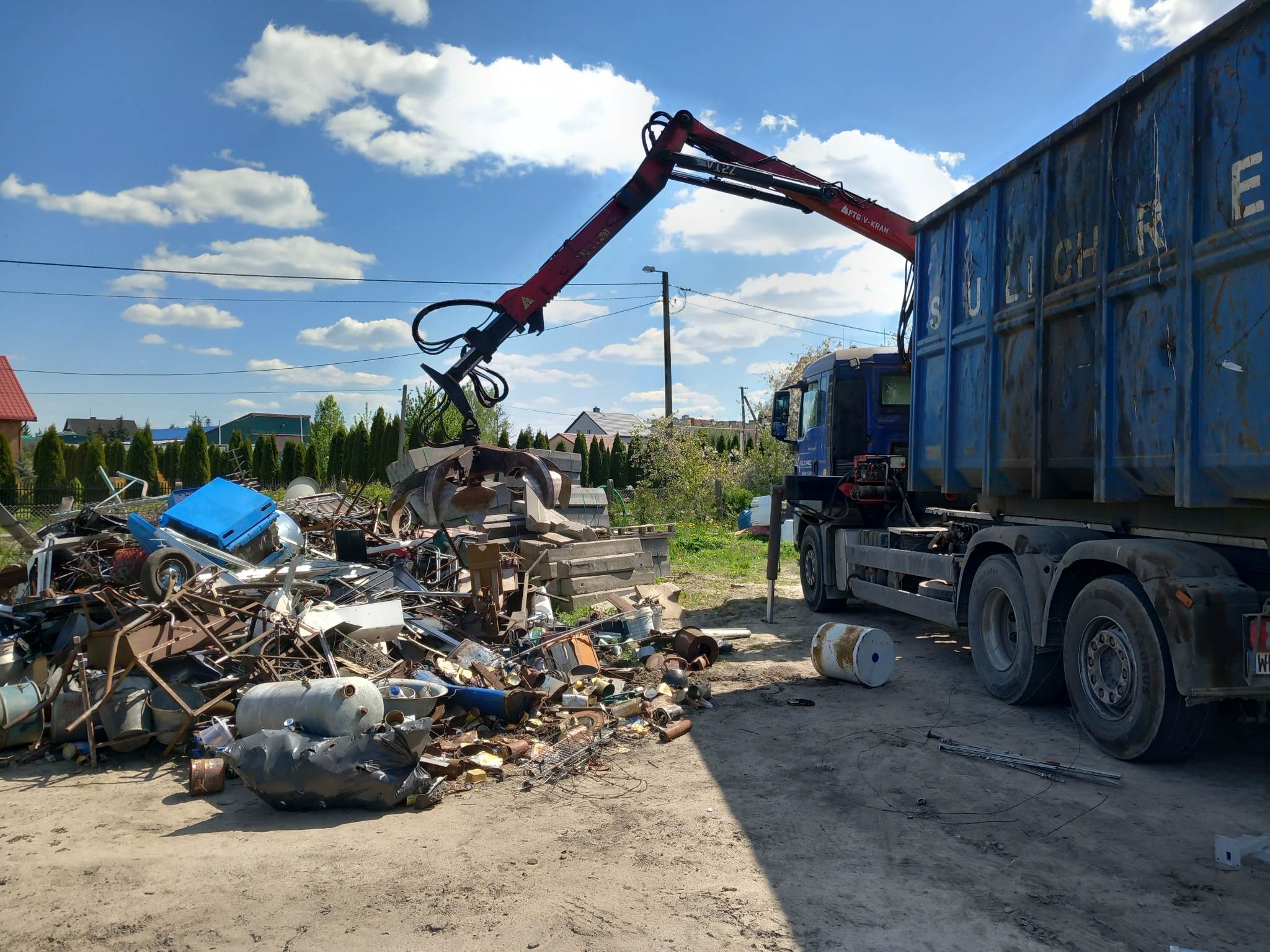 Dźwig mostowy podnosi metalowe odpady z montażu złomu na otwartej przestrzeni. W tle widać samochód ciężki z przyczepą wypełnioną gruzem i metalowymi odpadami oraz niebo z chmurami.