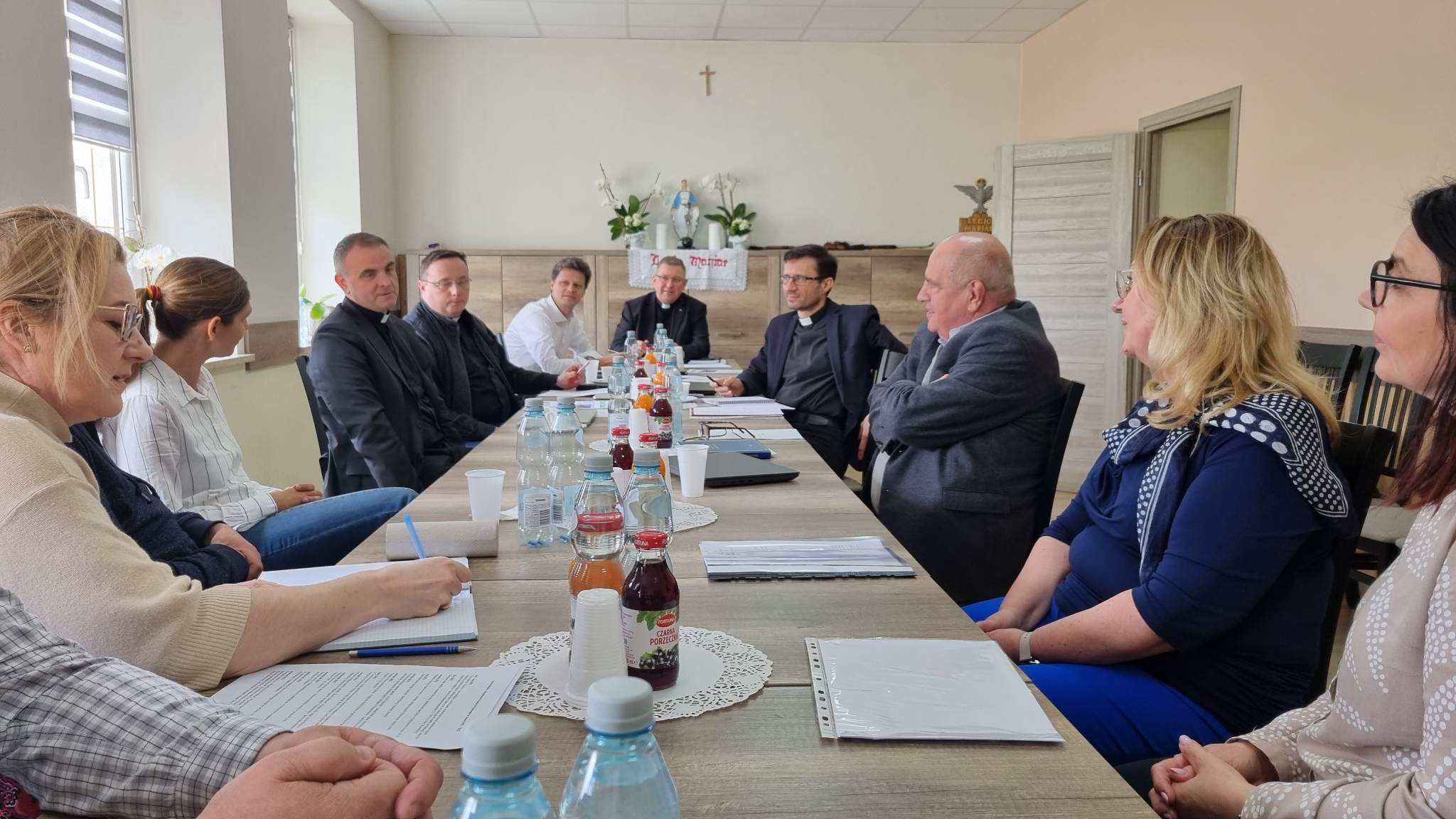 Na zdjęciu widać okrągły stół, wokół którego siedzi grupa osób. Wszyscy są ubranymi na co dzień, w różnych kolorach i fasonach. Na stole leżą dokumenty i napoje. Tło to jasna ściana z oknem.