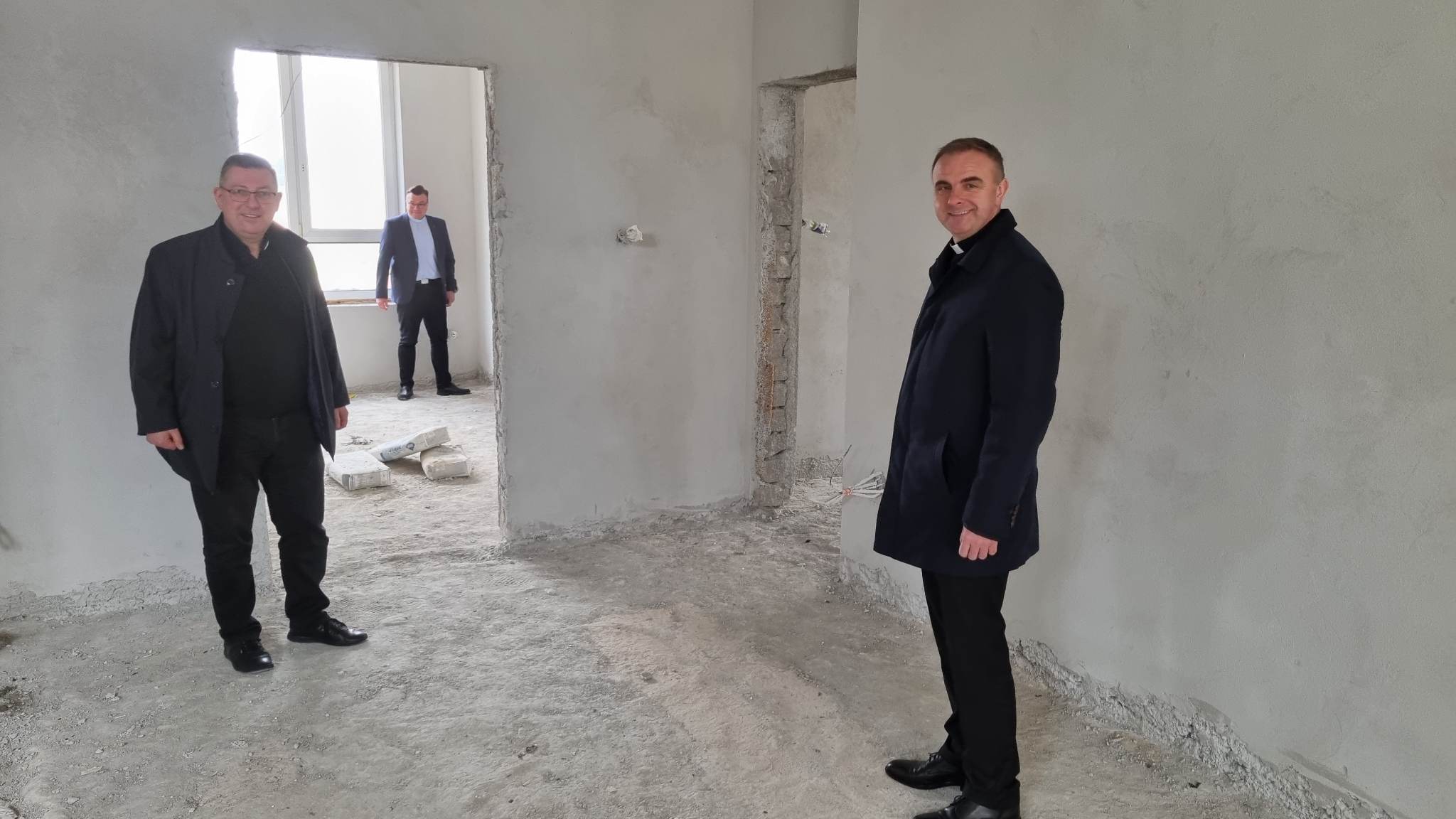 Dwoje osób w ciemnych marynarkach stoi w pustej, nieymalowanej przestrzeni. W tle widoczna jest ściana z cegieł i okno.