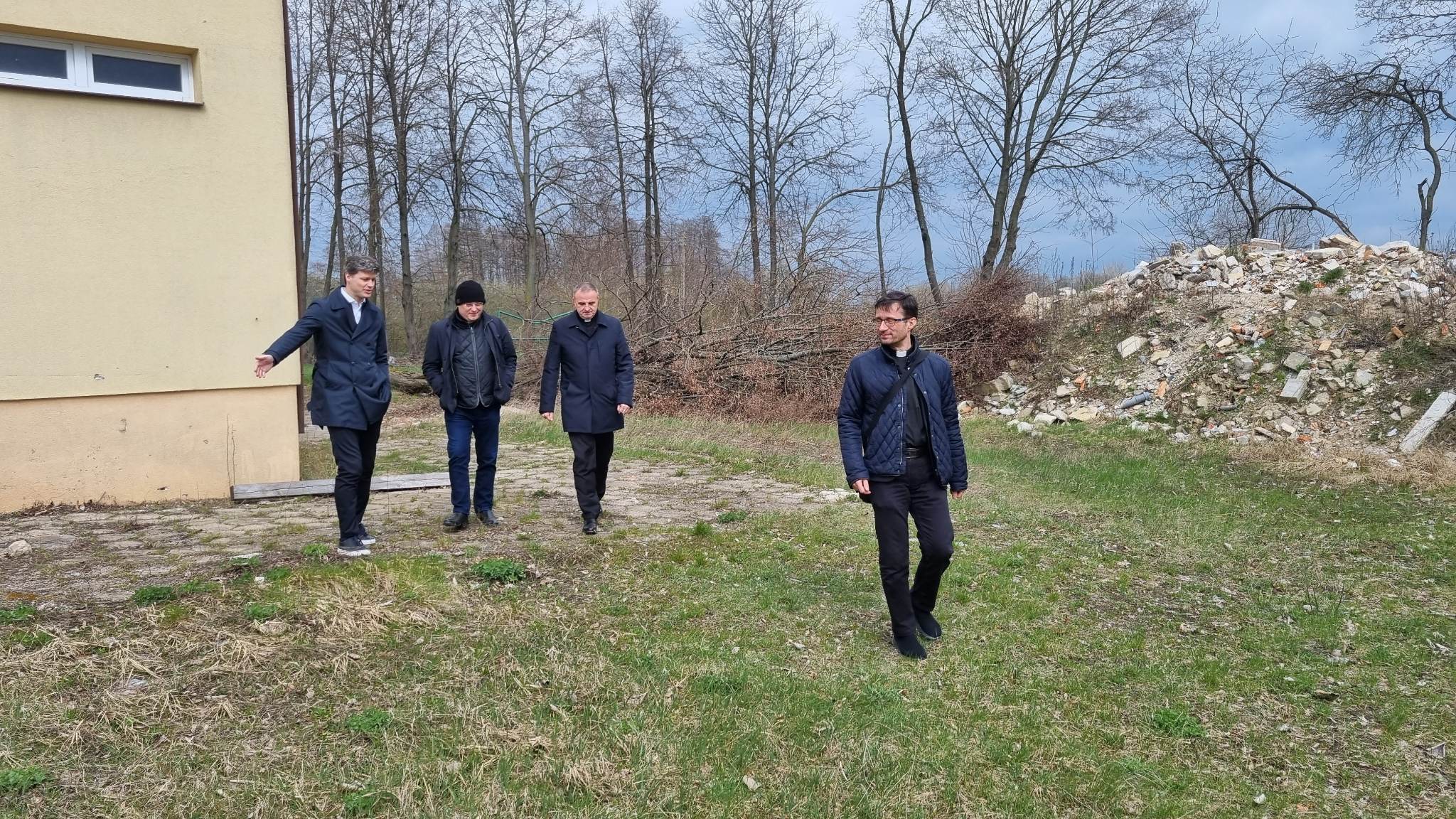 Cztery osoby w ciemnych płaszczach stoją na trawiastym terenie. W tle znajduje się budynek o pomarańczowej elewacji i drzewa. Na pierwszym planie stoi osoba z wyciągniętymi rękami. Zdjęcie przedstawia wizytę w Hodyszewie.
