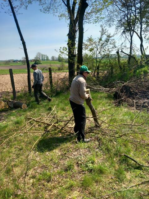 Mężczyźni sprzątają teren wokół domu. Jeden z nich trzyma gałązkę, a drugi idzie w oddali.