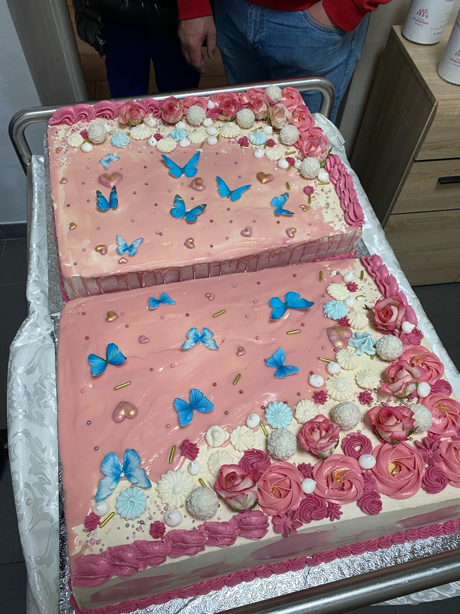 Duży, różowy tort udekorowany kwiatami i motylami, prezentowany na aluminiowej tacy. W tle drewniana szafka z pojemnikami.