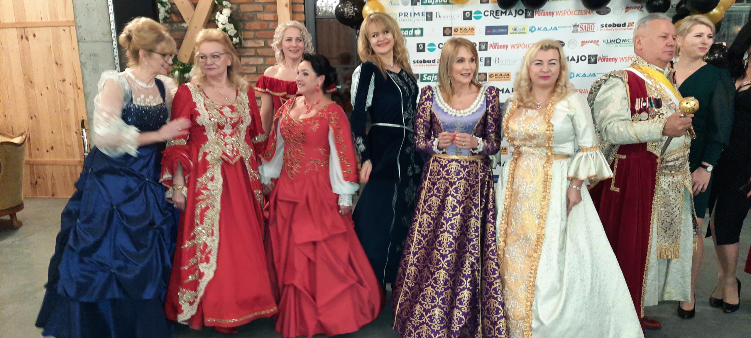 Grupa osób w strojach nawiązujących do epoki staropolskiej stoi w rzędzie. Widać czerwone, czarne i brązowe płaszcze oraz sukienki z koronkowymi detalami. Na tle znajduje się ciemne tło z reklamami.
