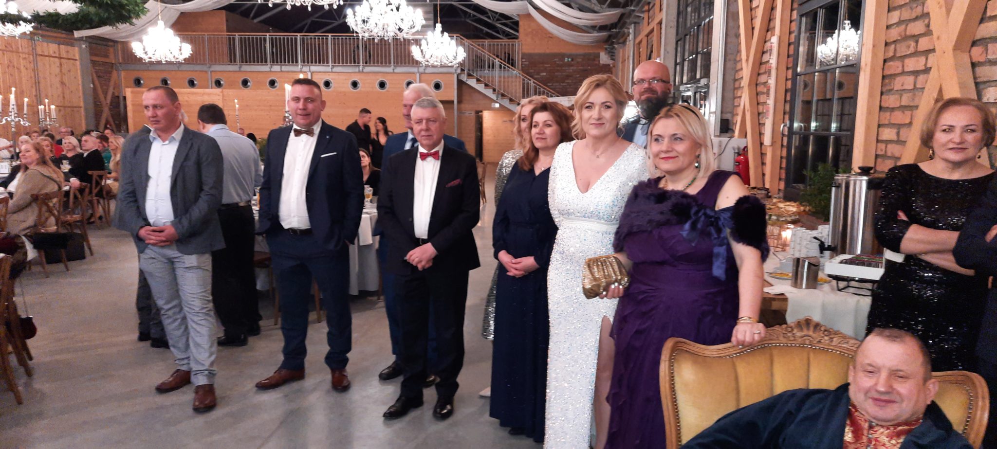 Na zdjęciu przedstawiono grupę osób w eleganckich strojach na balu charytatywnym. W tle znajduje się drewniany salon z żyrandolami i stołem z drinkami. Uczestnicy są ubrani w formalne stroje, w tym marynarki i suknie.