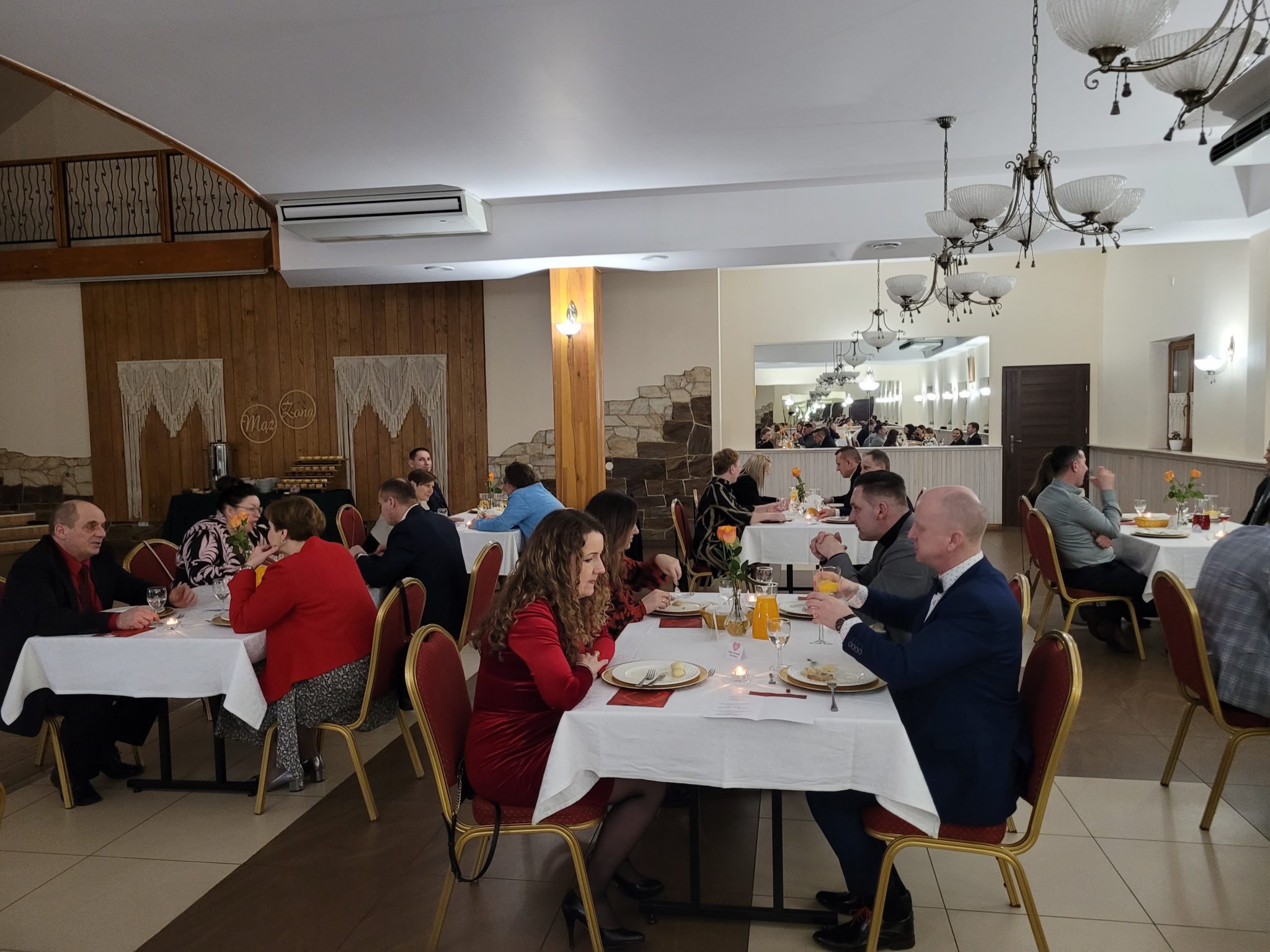 Wnętrze restauracji, w której odbywa się Małżeński Wieczór Walentynkowy. Na stolikach znajdują się talerze i szklanki, a osoby spożywają posiłek. W tle znajduje się dekoracja ścienna z kamiennym tłem. Uczestnicy są ubrani na różne sposoby.