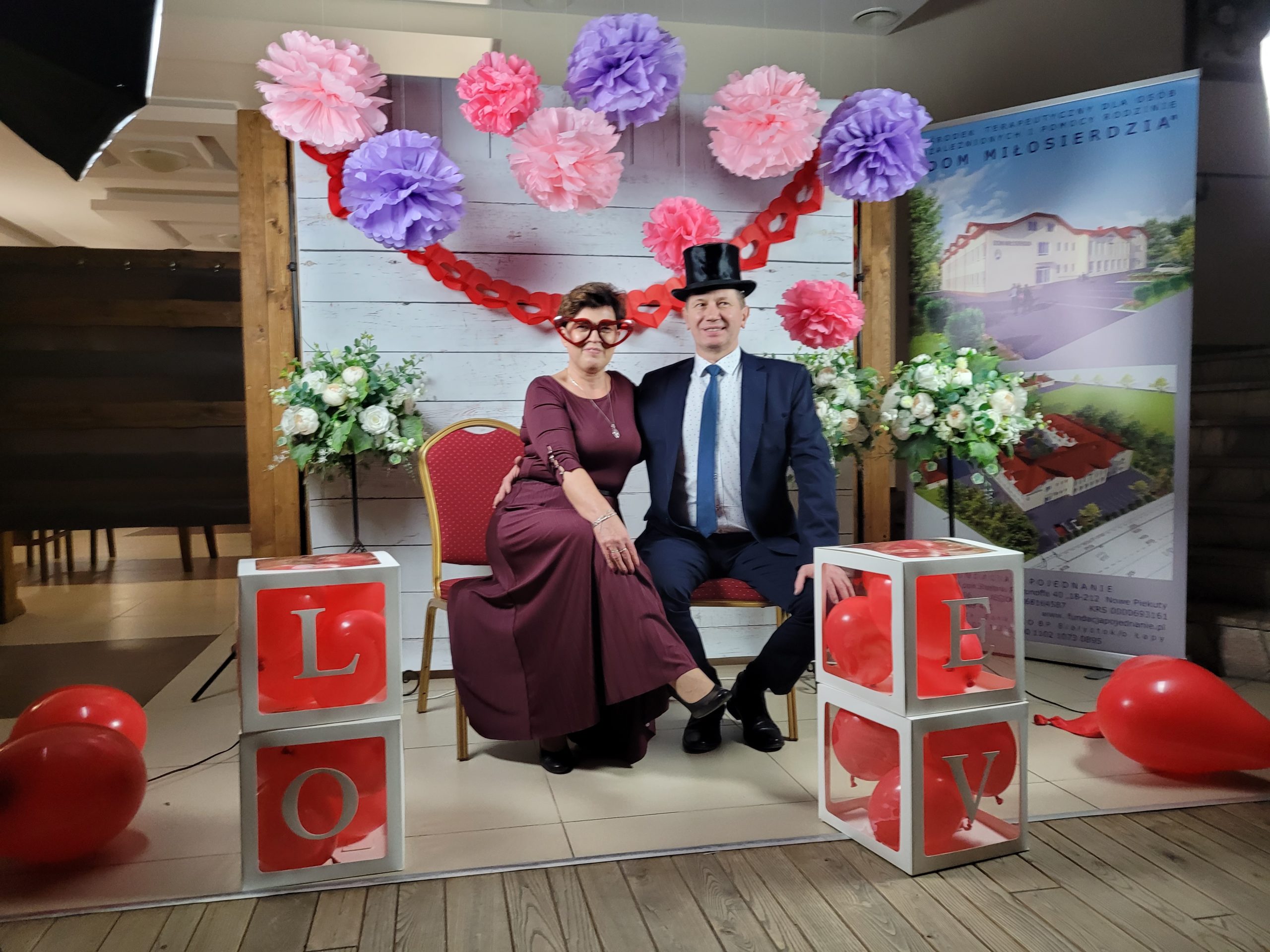 Na zdjęciu dwoje osób w formalnych strojach siedzi przy drewnianych blokach z literami „L” i „X”. W tle znajduje się dekoracja walentynkowa i plakat przedstawiający dom.