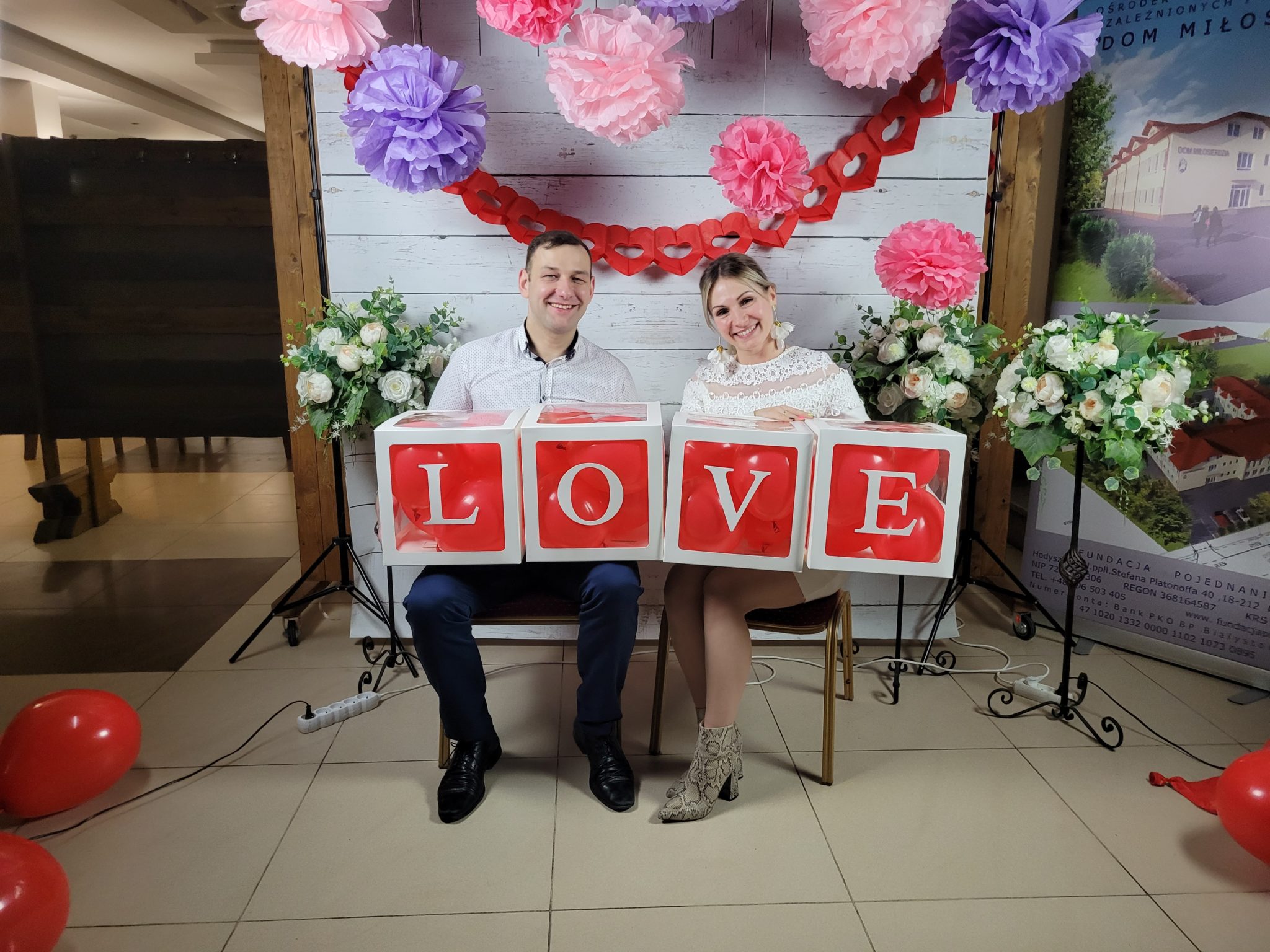 Para siedzi na krześle i ławce przed dużym ekranem z literami 'LOVE' w czerwonym kolorze. W tle znajduje się dekoracja z pomponami, kwiatami i obrazkiem krajobrazowym. Na podłodze leżącymi kulkami.