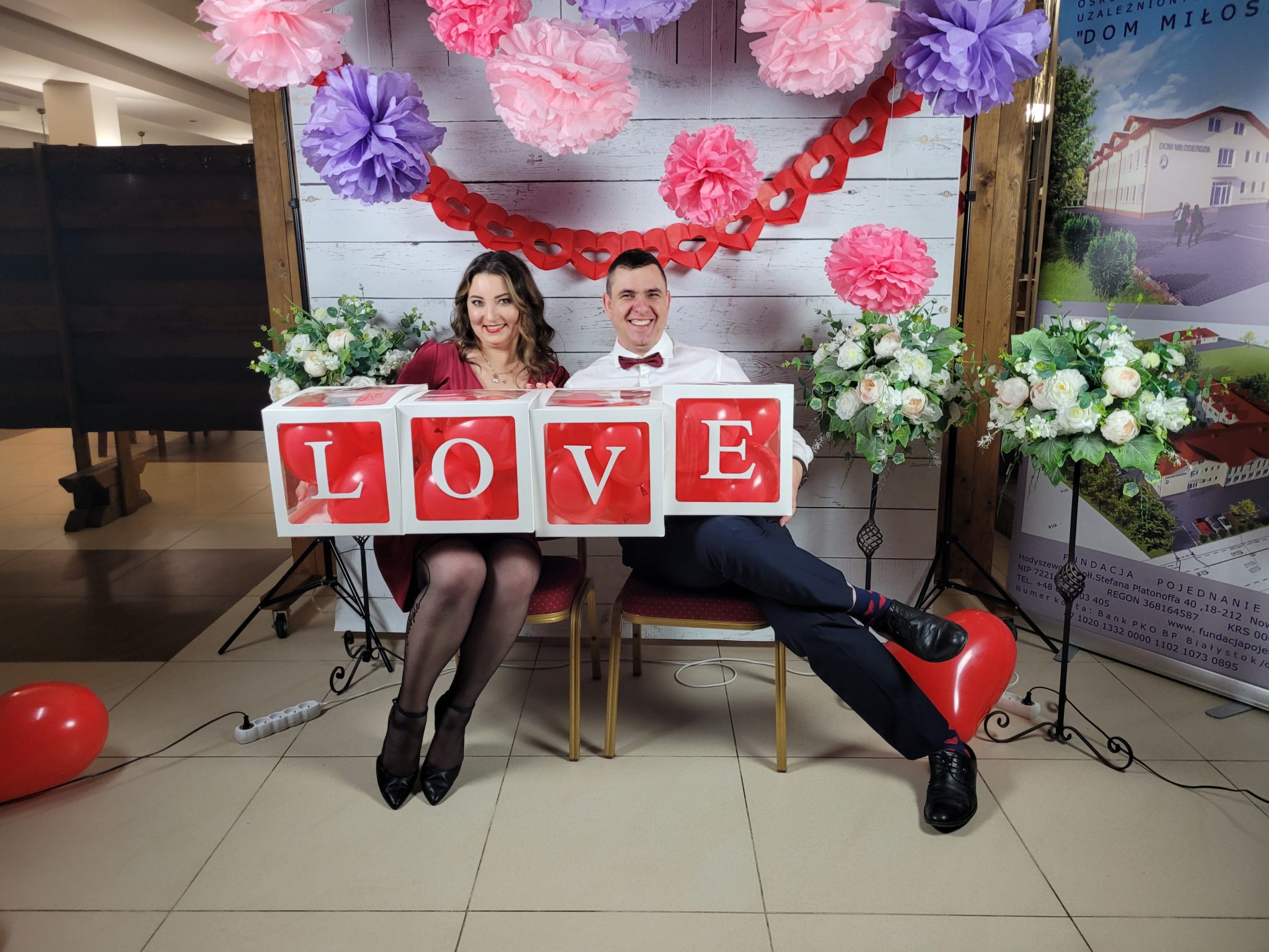 Para siedzi przed tłem z dekoracjami walentynkowymi i trzyma w rękach litery tworzące słowo "LOVE". Na zdjęciu widoczny element wystawy podczas wydarzenia "Małżeński Wieczór Walentynkowy".