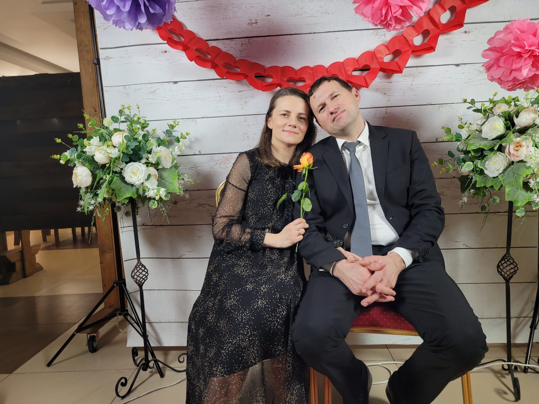 Para w formalnych strojach uśmiecha się do kamery na tle dekoracyjnej ściany z pomponami i sercami. Na zdjęciu widoczny jest również bukiet kwiatów.