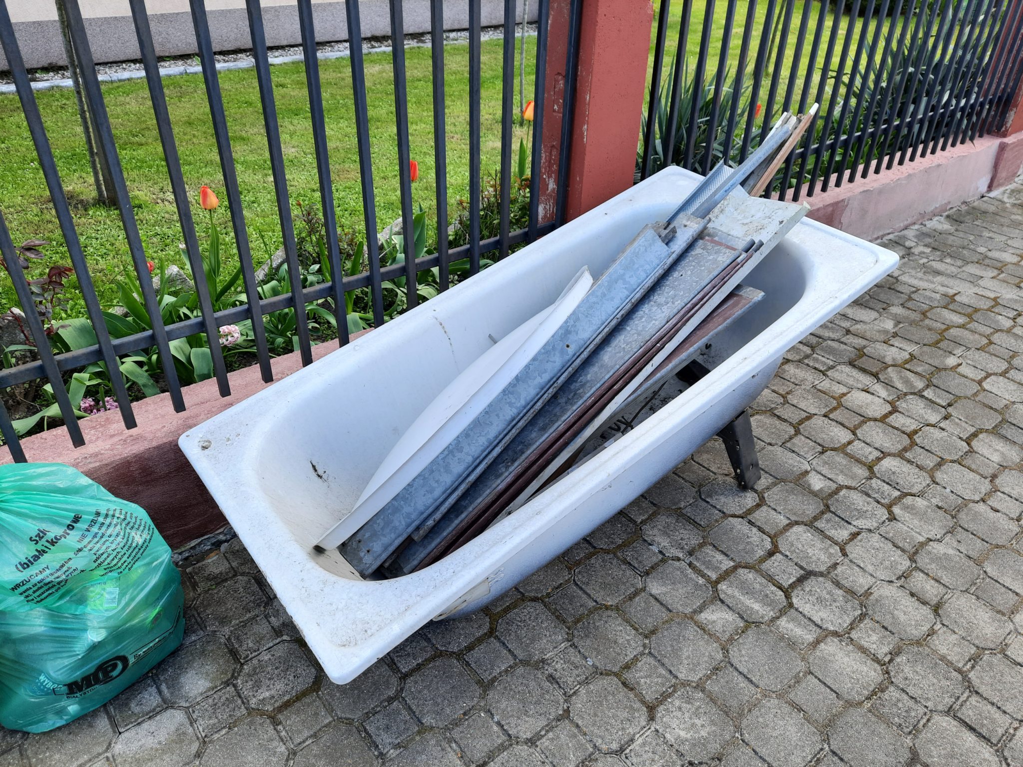 Na zdjęciu widoczna stara wanna z metalowymi elementami leżąca na chodniku w ogrodzie. W tle znajduje się płot i roślinność. Worek foliowy stoi po lewej stronie.