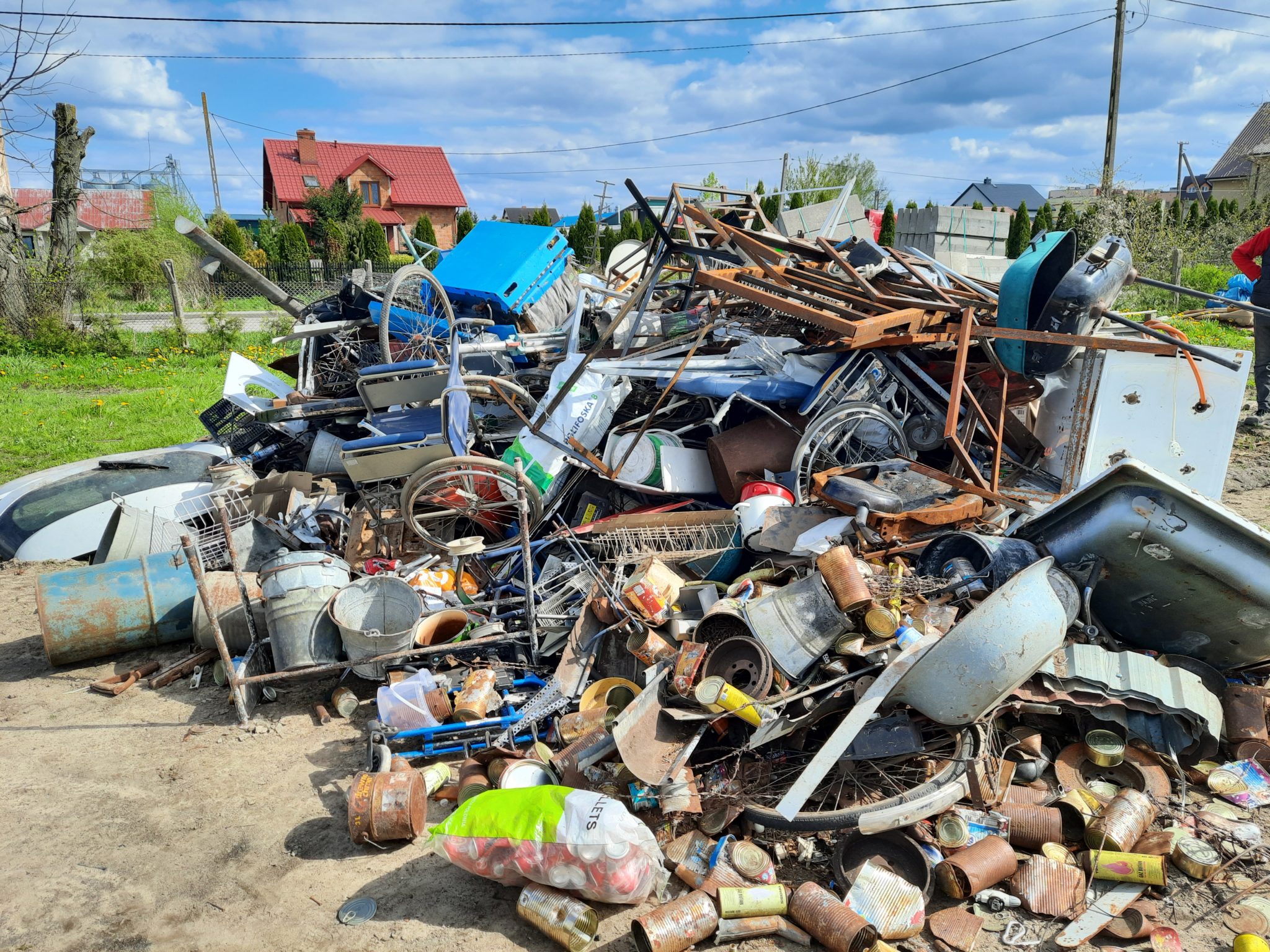 Zbierka złomu na otwartej przestrzeni, składająca się z różnych elementów metalowych i plastikowych. Na zdjęciu widać stos odpadów na tle domu i nieba.
