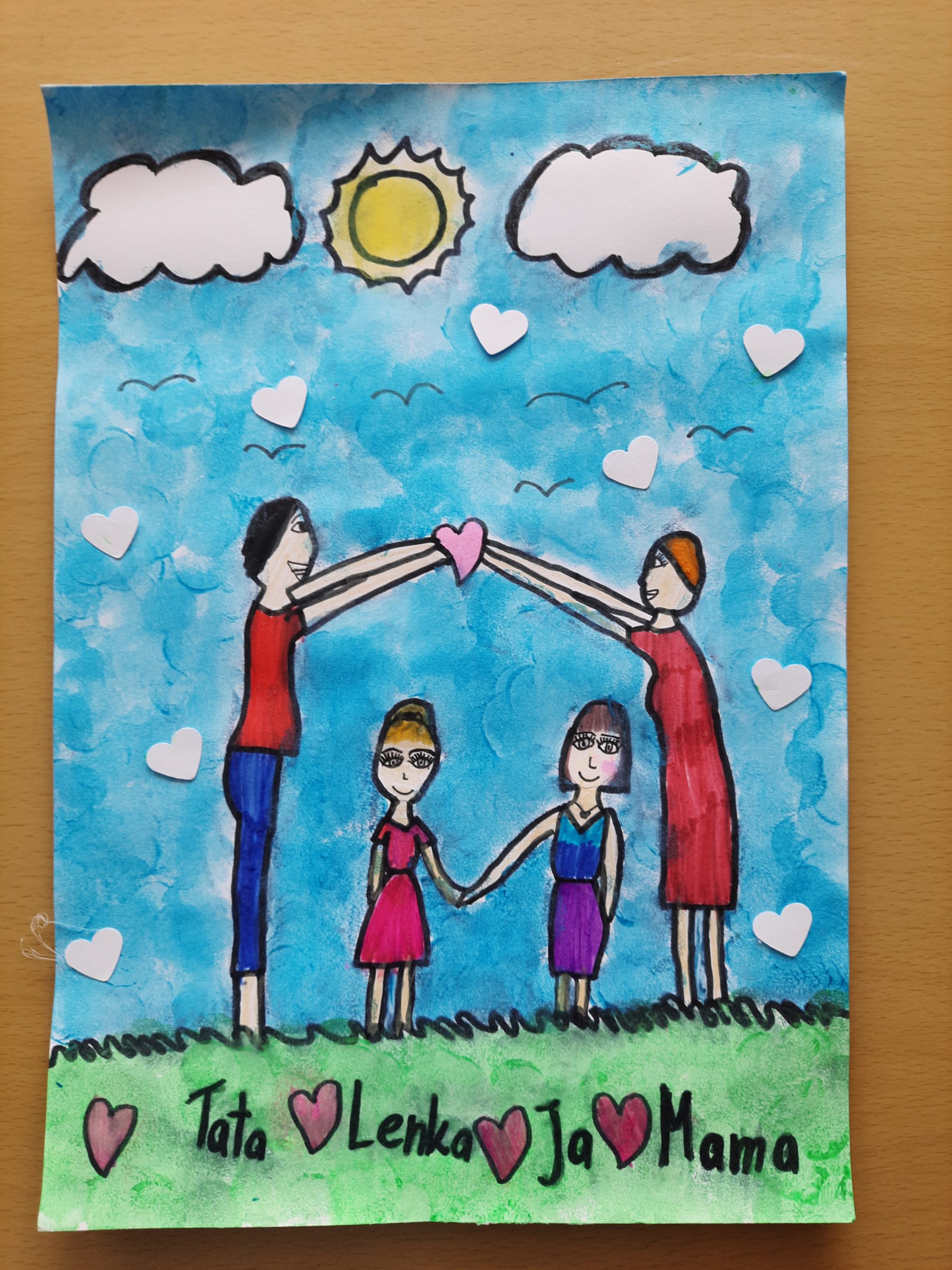 Rysunek przedstawiający rodzinę w kształcie domu zbudowanego z serc. Na niebie są słońce i chmury. Pod obrazkiem napisane jest „Tata ❤️Lenka❤️Ja❤️Mama”.
