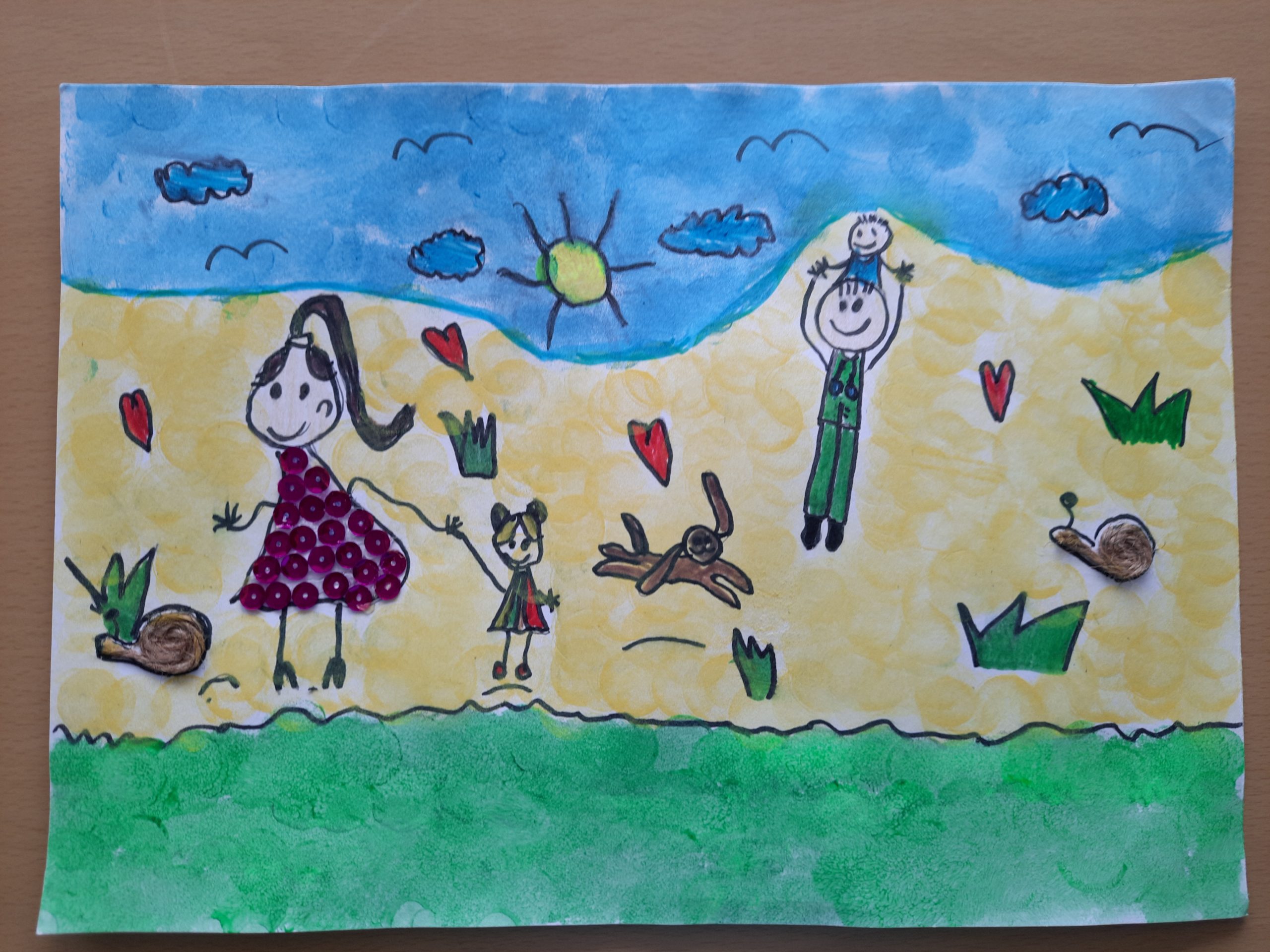 Ilustracja przedstawiająca rodzinę na plaży, z dziećmi bawiącymi się i dorosłą osobą wznoszącą się w powietrze. Na obrazku widoczny jest pies oraz słońce i chmury.
