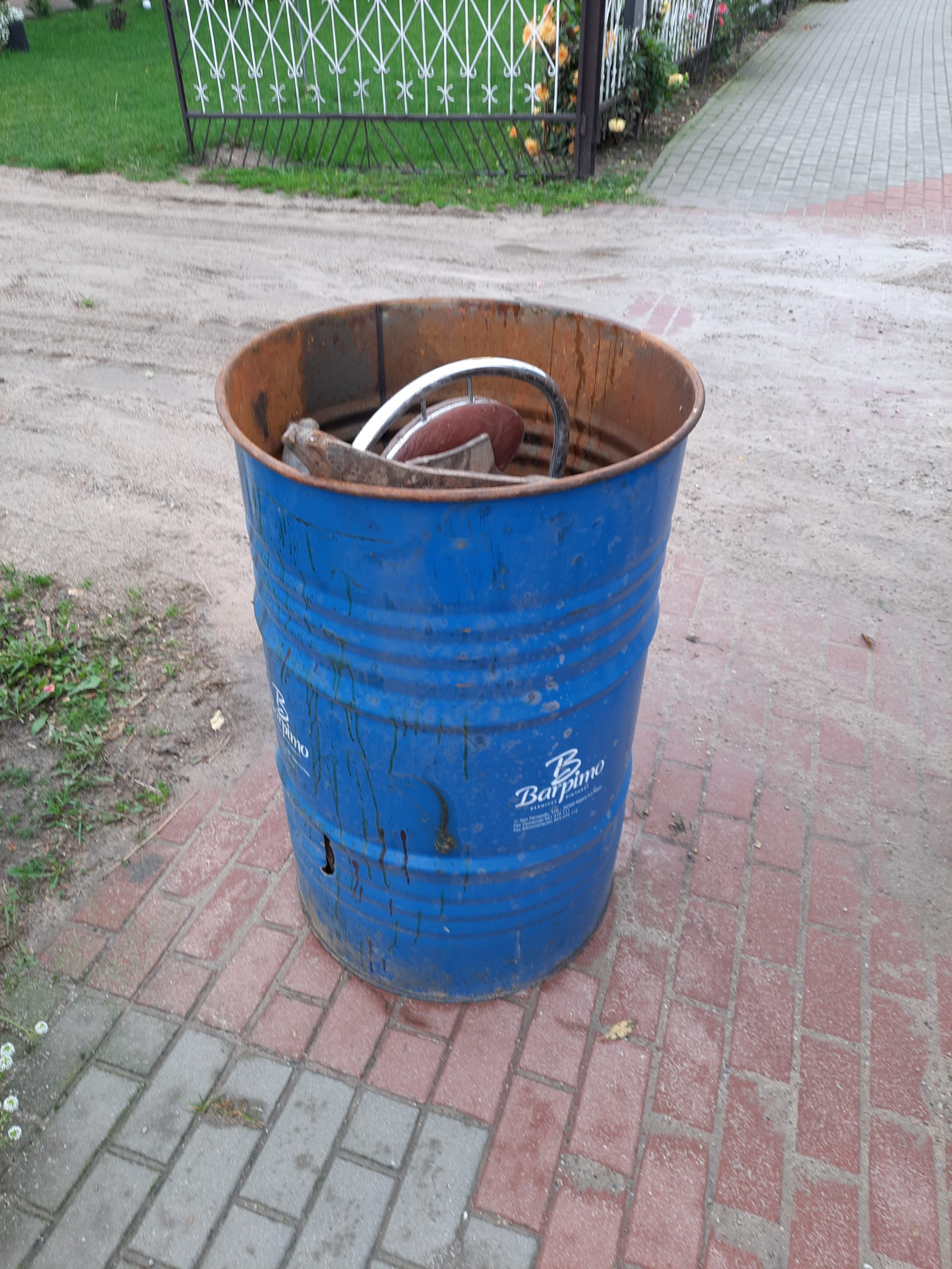 Zdjęcie kosza na śmieci wypełnionego metalową rurą i ciemnym materiałem, znajdującego się na chodniku obok trawnika. Na tle widać element ogrodzenia z siatki.