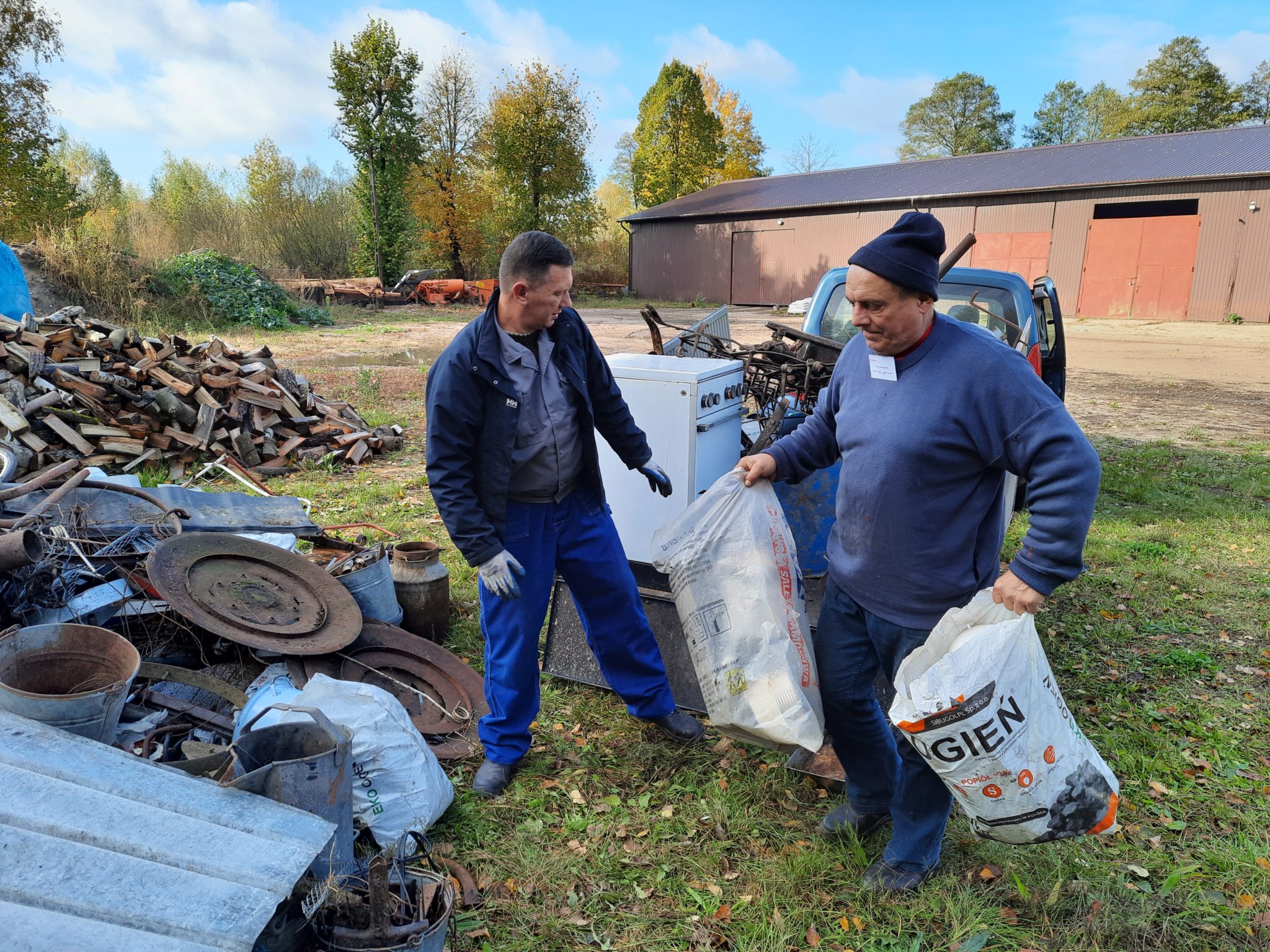 Dwoje osób stoi na trawniku przed budynkiem. W tle widać drewniane konstrukcje, paletę drewna i samochód. Jedna osoba trzyma duży worek foliowy, a druga ma w ręku urządzenie elektryczne.
