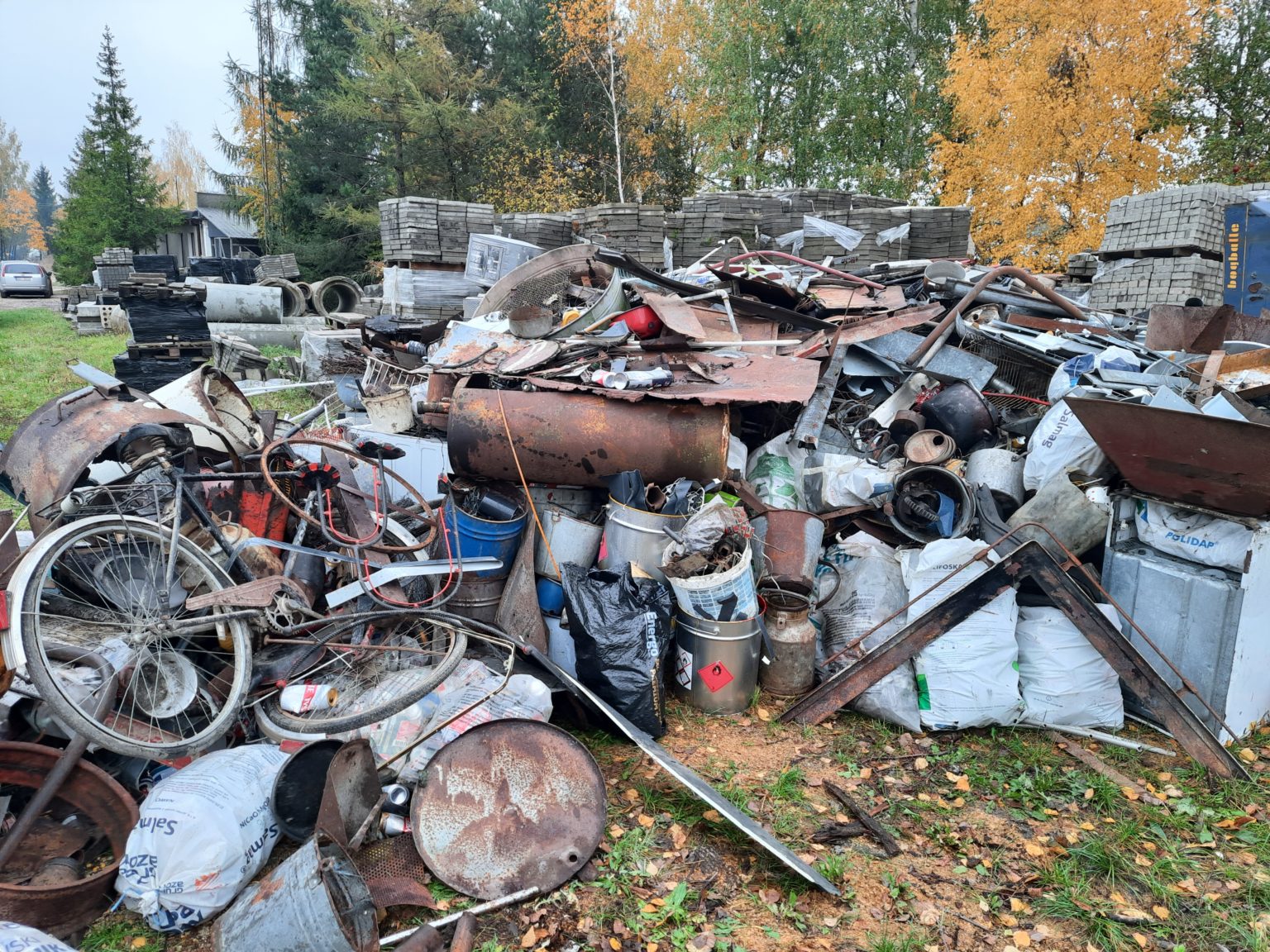 Na stercie leżą metalowe elementy, części rowerowe, worki i pojemniki. W tle zielony krajobraz z drzewami i budynkami. Zbiórka złomu w gminie Brańsk zakończona.