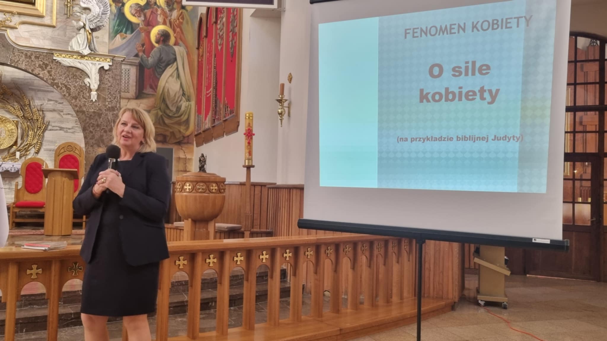 Na projekcyjnym ekranie widnieje tekst "FENOMEN KOBIE TY O SILE KOBIE TY (na przykładzie biblijnej Judyty)". Prezentacja dotyczy tematu siły kobiety, z wykorzystaniem postaci biblijnej Judyty.
