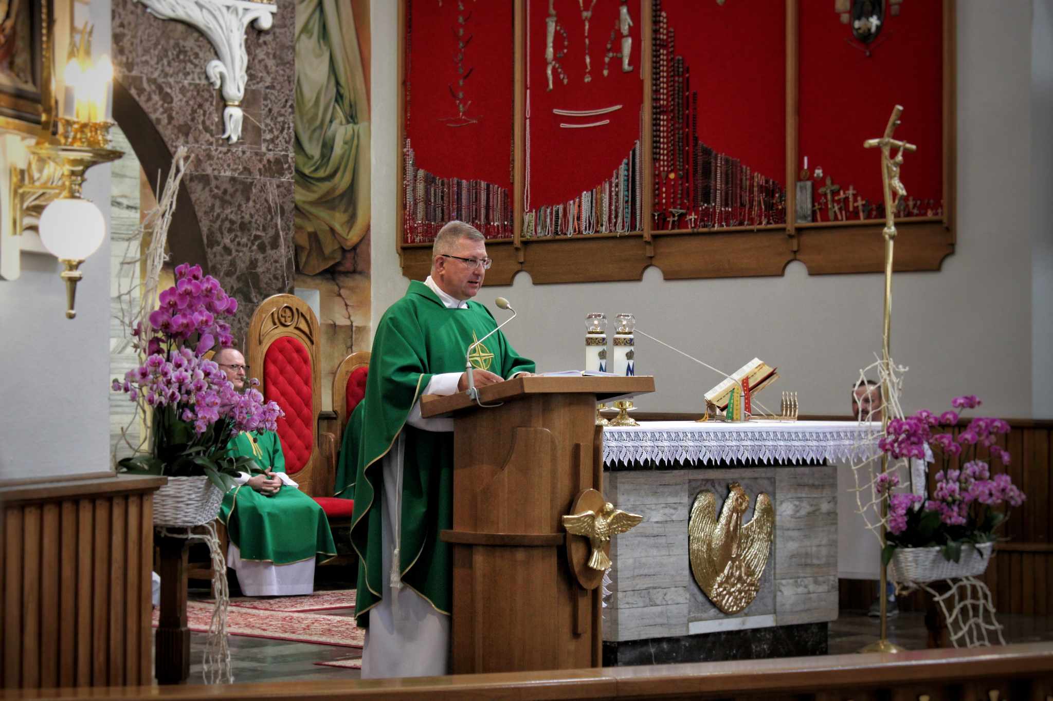 Mężczyzna w stroju celebracyjnym prowadzi uroczystość eucharystyczną w kościele, otoczony kwiatami i innymi uczestnikami. Na ścianach znajdują się obrazy. Widać ołtarz z elementami dekoracyjnymi.