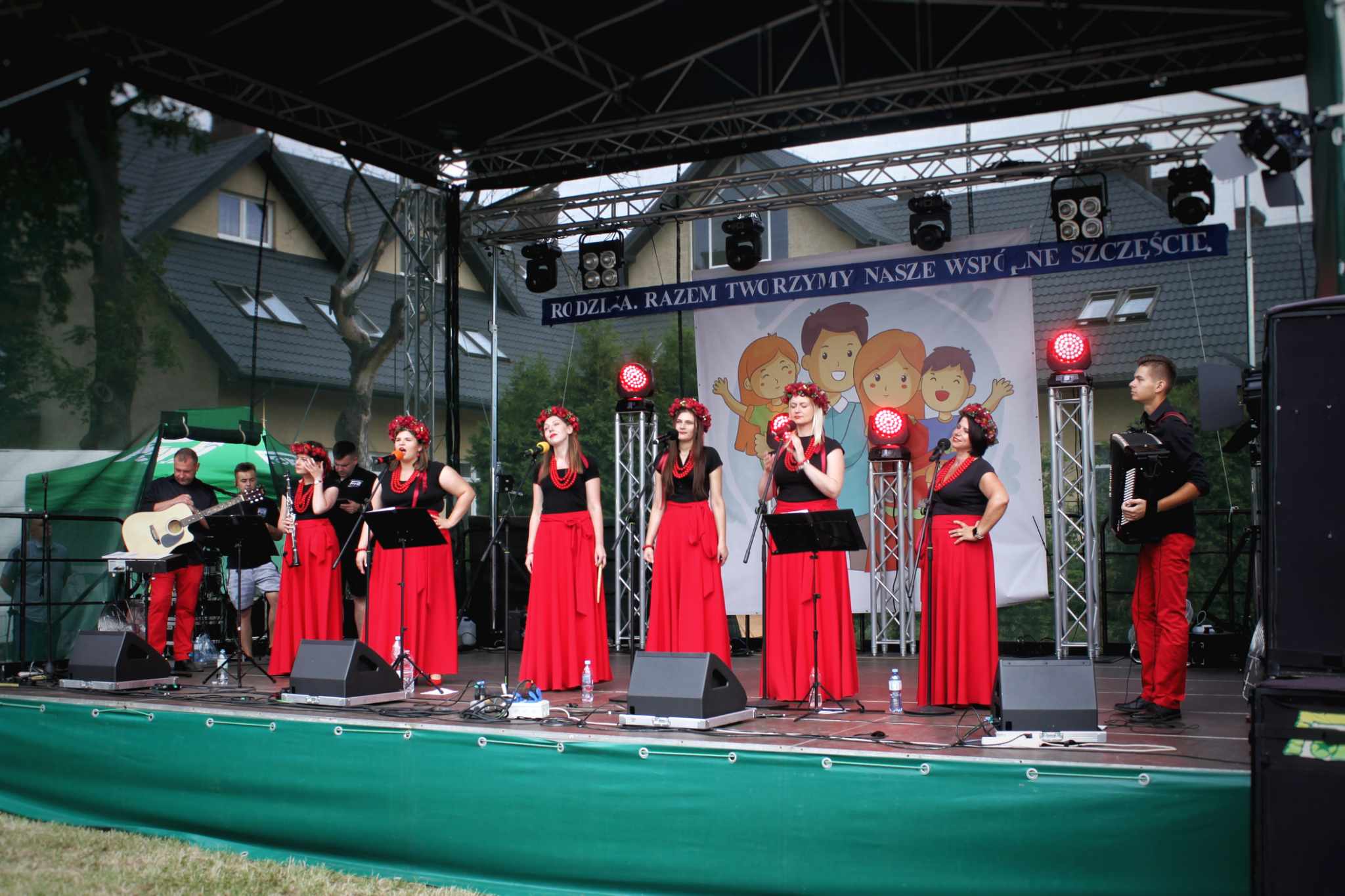 Grupa wokalistów i muzyków w czerwonych strojach występuje na scenie podczas wydarzenia "Dzień Rodziny". Na plakatach widnieje napis „REMEMBER KOTY NASZE WSZYSTKIE”.