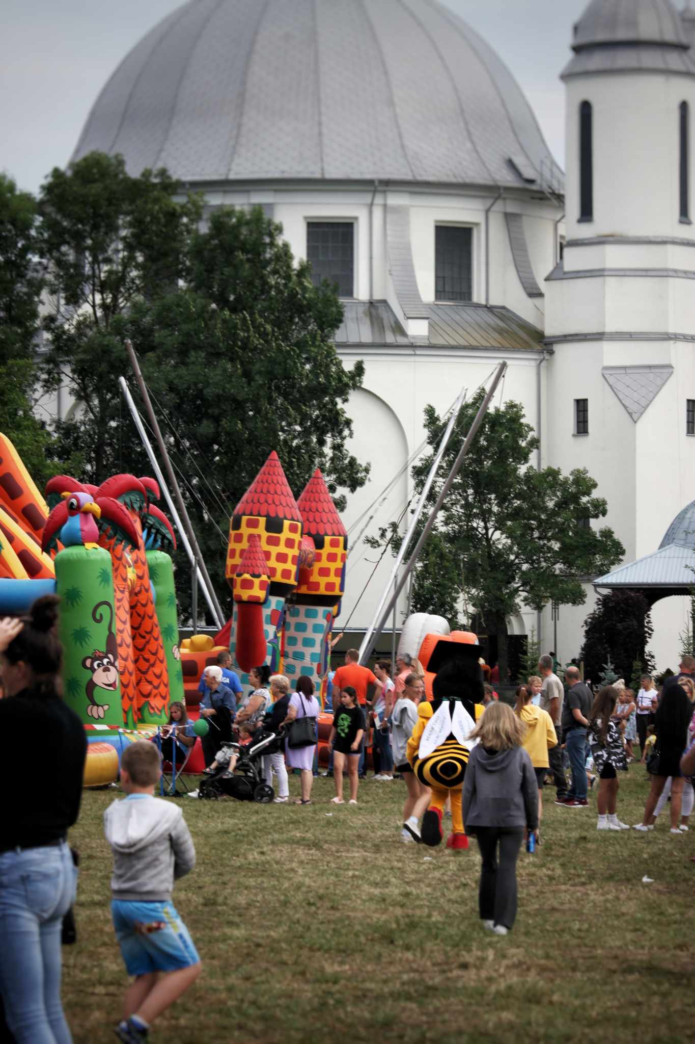Na placu festynowym widać dmuchane atrakcje, dzieci bawiące się i grupę ludzi. W tle znajduje się kościół o kopułach. Użyte elementy to: zamek, zjeżdżalnia, dmuchane bańki, drzewa, budowla kościelna.