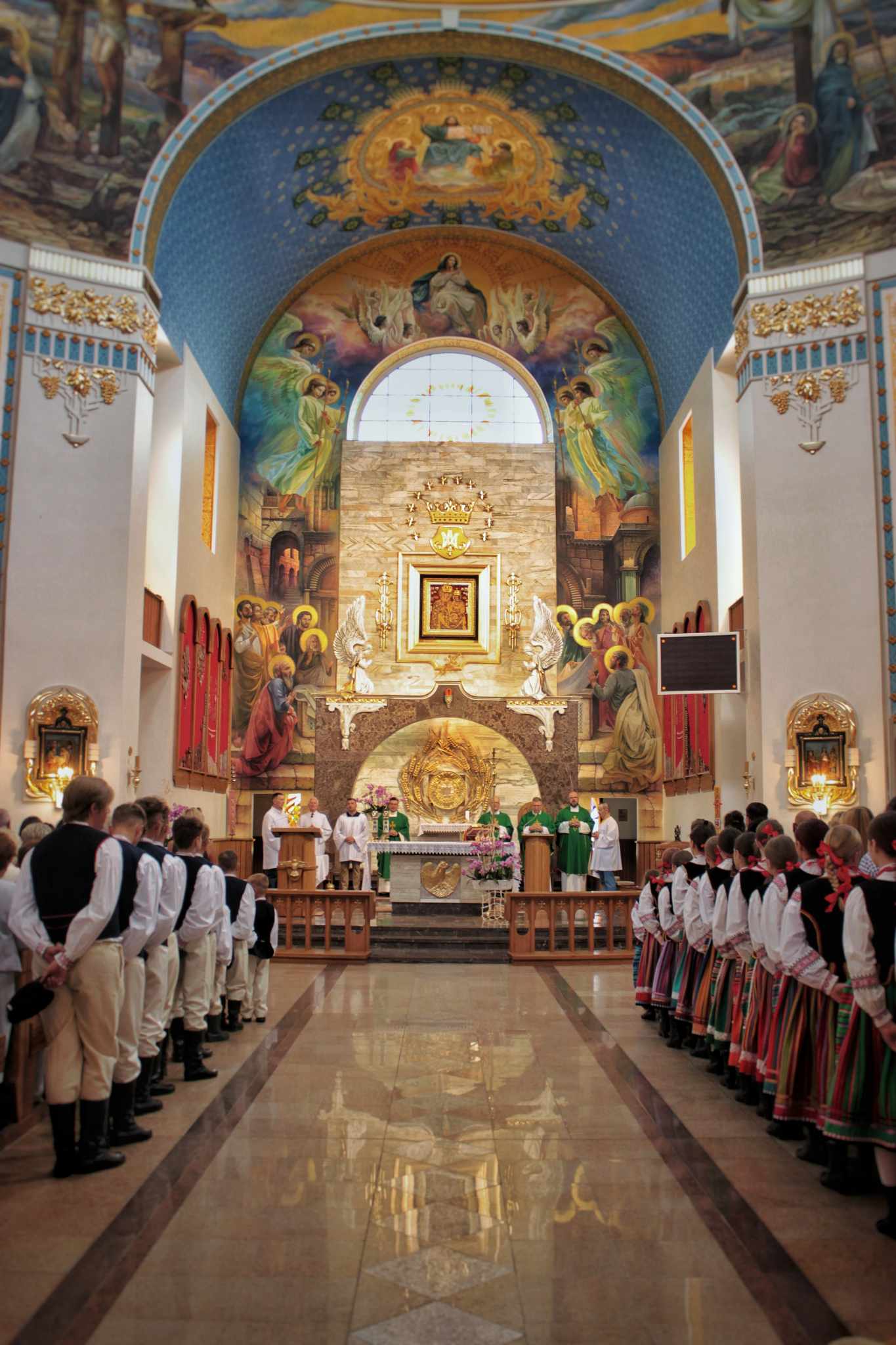 W kościele odbywa się uroczysta Eucharystia. Grupa osób w strojach ludowych stoi w rzędach po obu stronach ołtarza, a na ścianie znajduje się duży obraz przedstawiający scenę religijną.