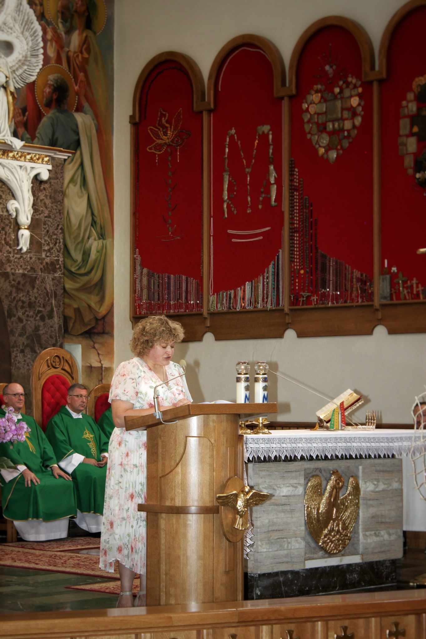 Wnętrze kościoła podczas uroczystości eucharystycznej. Na platformie stoi osoba trzymająca mikrofon, a w tle siedzą księża i ołtarz z kieliszkami.