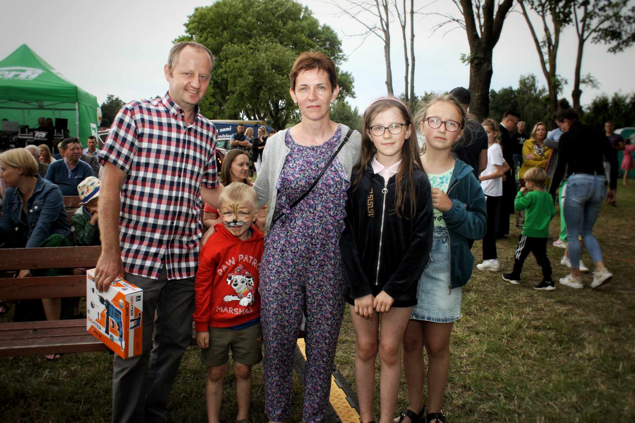 Zdjęcie przedstawia rodzinę na pikniku. Widać mężczyznę w koszuli w kratę, kobietę w kombinezonie oraz dwie córki w okularach. Na pierwszym planie trzyma pudełko dziecko. W tle znajdują się inni ludzie i elementy krajobrazu.
