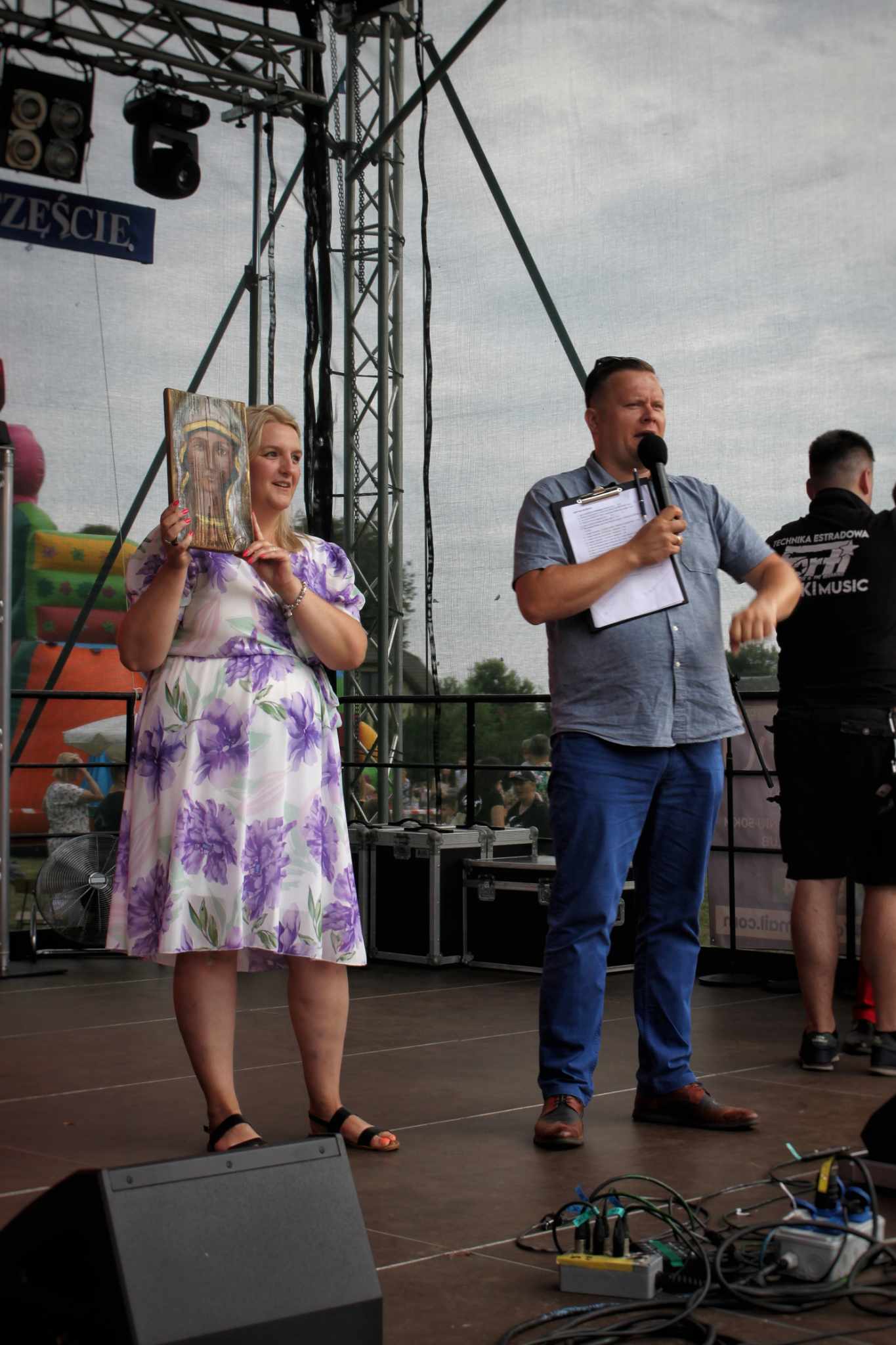 Dwie osoby stojące na scenie podczas wydarzenia, osoba po lewej trzyma książkę, a osoba po prawej ma notatki. W tle widoczna konstrukcja sceniczna i dekoracje.