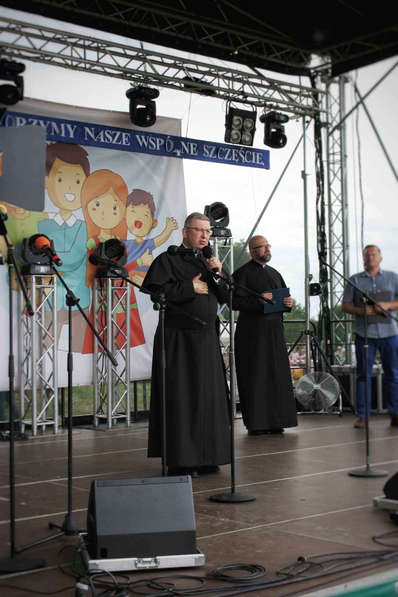 Kapłan przemawia do mikrofonu na scenie podczas wydarzenia rodzinnego w Hodyszewie; za nim stoi drugi duchowny.