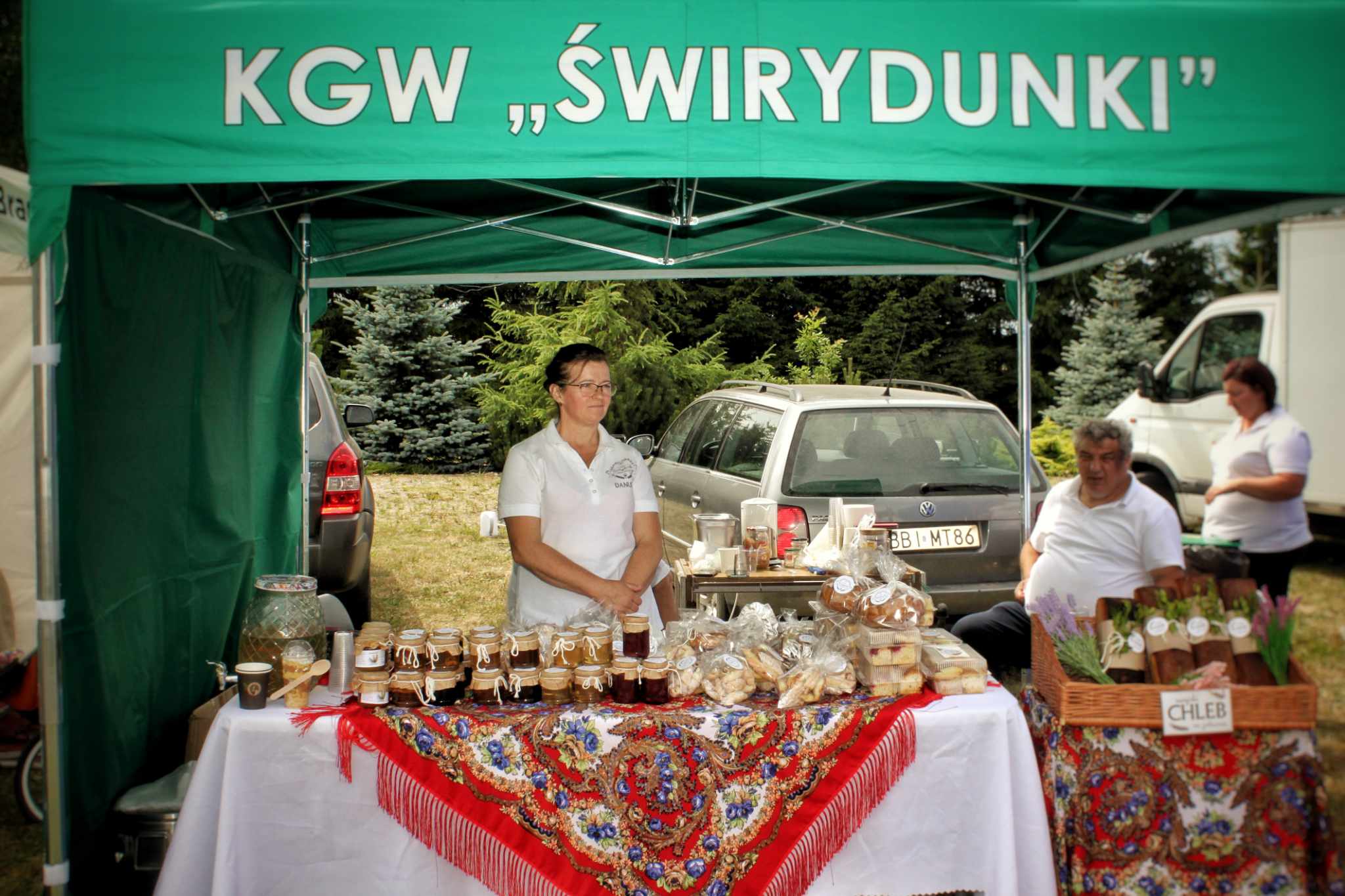 Na stoisku KGW „Świrydunki” prezentowane są ręcznie robione produkty spożywcze, w tym sękacze i serniki. W tle znajduje się samochód oraz mężczyzna z koszykiem warzyw.