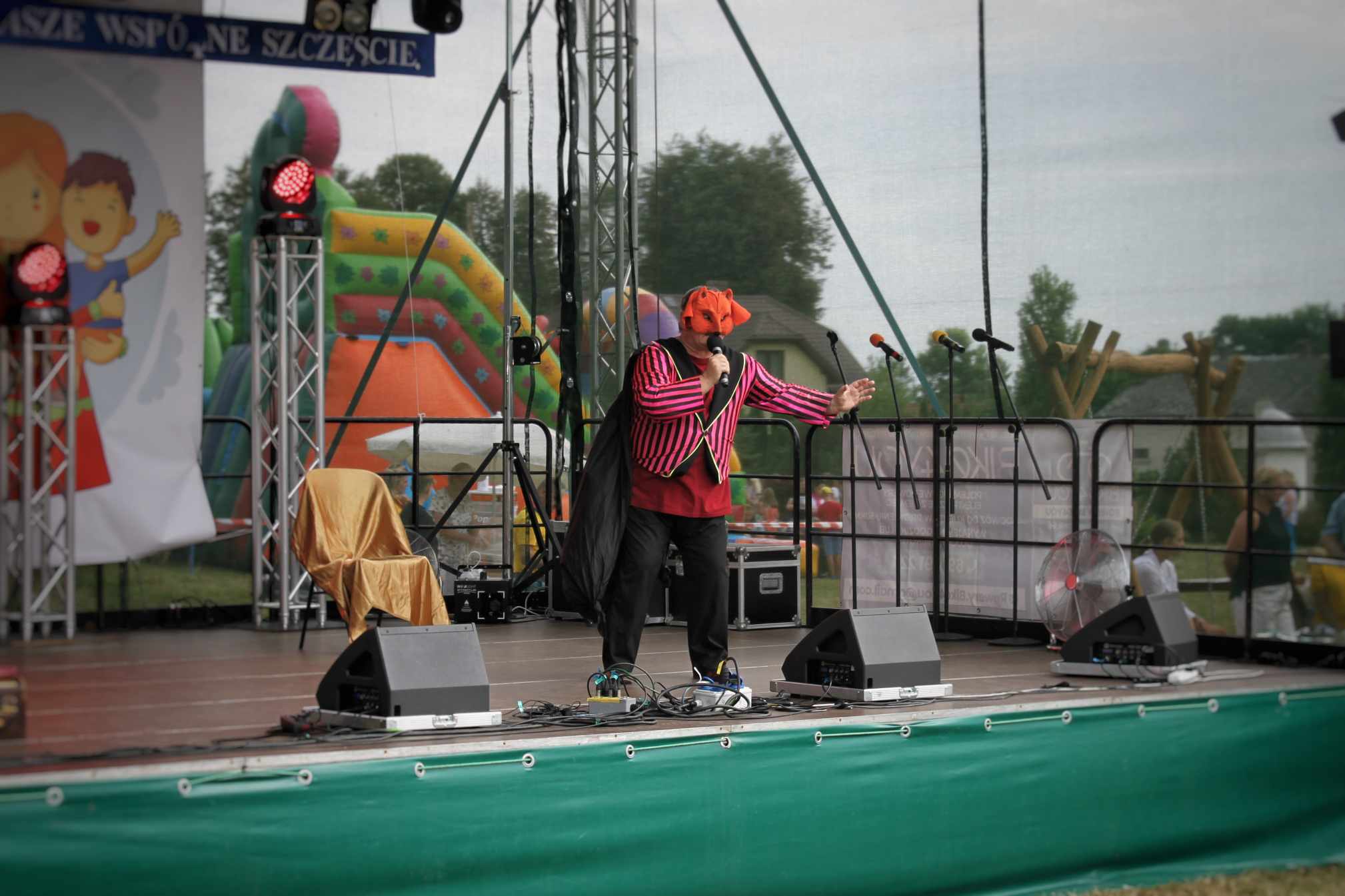 Na scenie występuje osoba w czerwono-czarnym stroju z pomarańczową opaską na głowie, trzymając instrument muzyczny. W tle znajduje się plac zabaw i baner 'W Dzień Rodziny'.