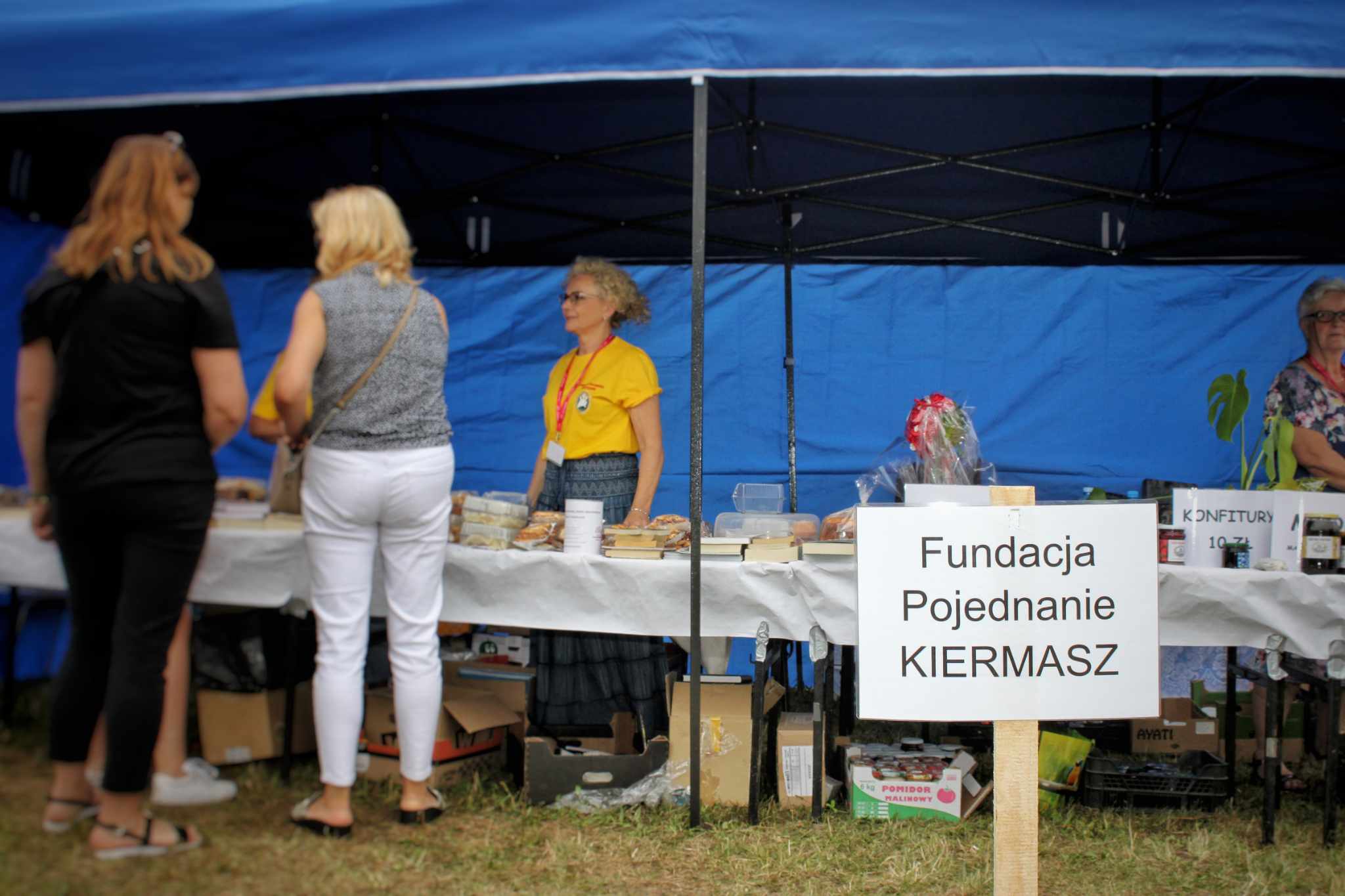 Na stoisku prezentowane są produkty, a wokół niego znajdują się osoby. Na płótnie widnieje napis „Fundacja Pojednanie KIERMASZ”.
