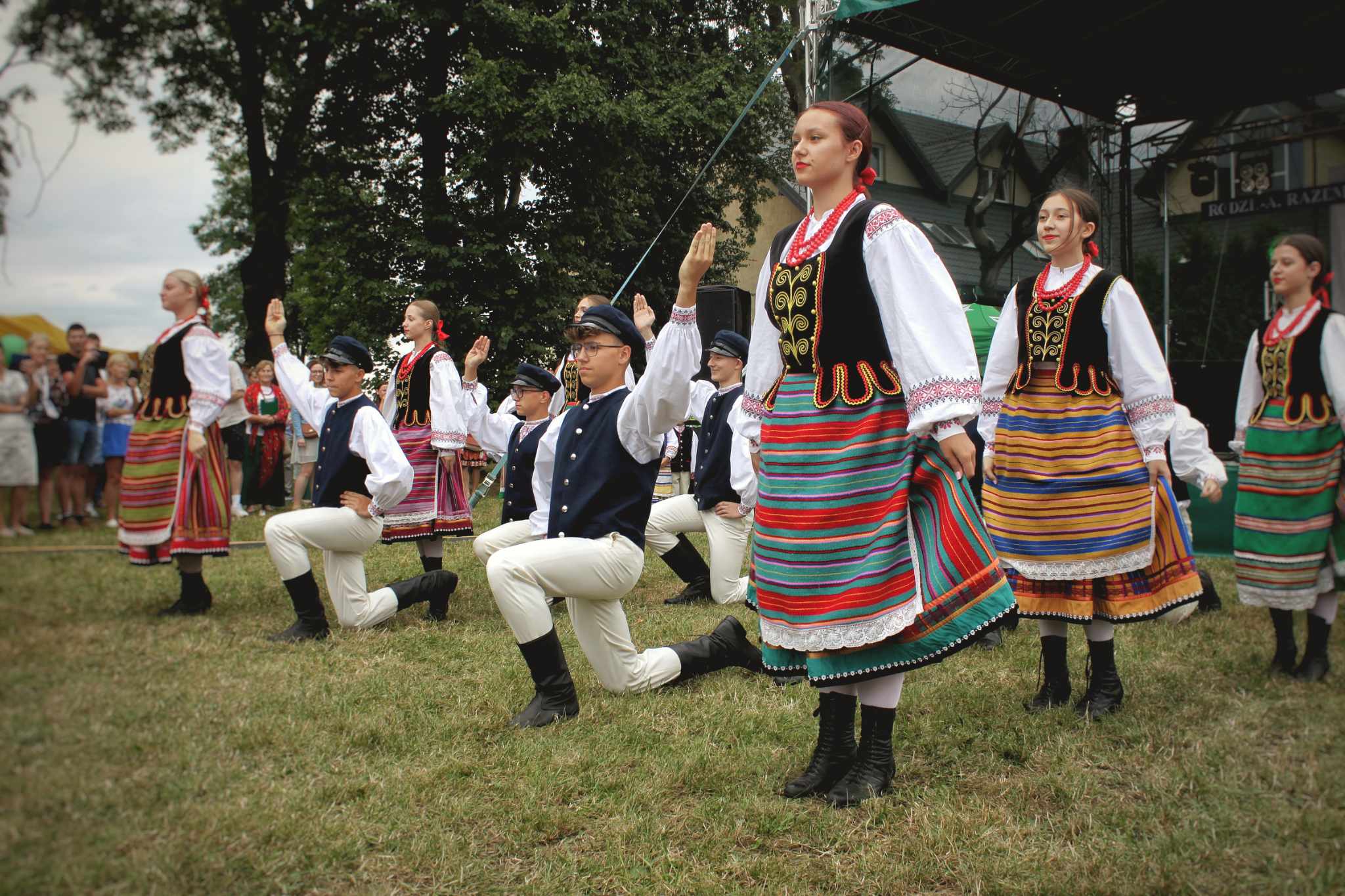 Grupa osób w strojach ludowych tańczy na otwartym terenie, wykonując rytuał. Widać kolorowe spódnice i tradycyjne elementy ubioru. Uczestnicy są ubranymi w białe koszule i spodnie.
