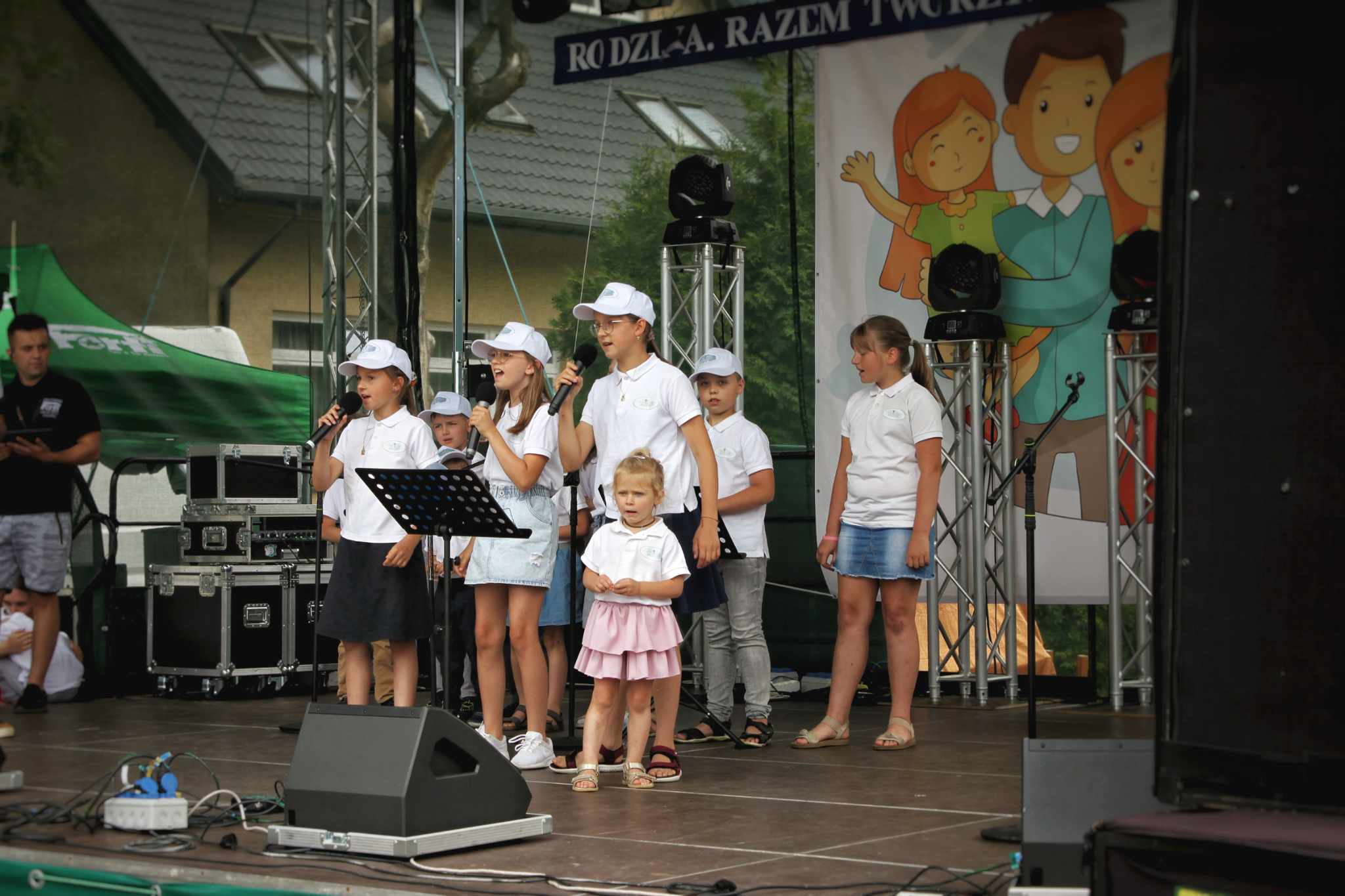 Na scenie w parku grupa dzieci w białych koszulach i czarnych beretach śpiewa. Wokół sprzęt nagłośnieniowy, publiczność i plakat z ilustracją rodziny.