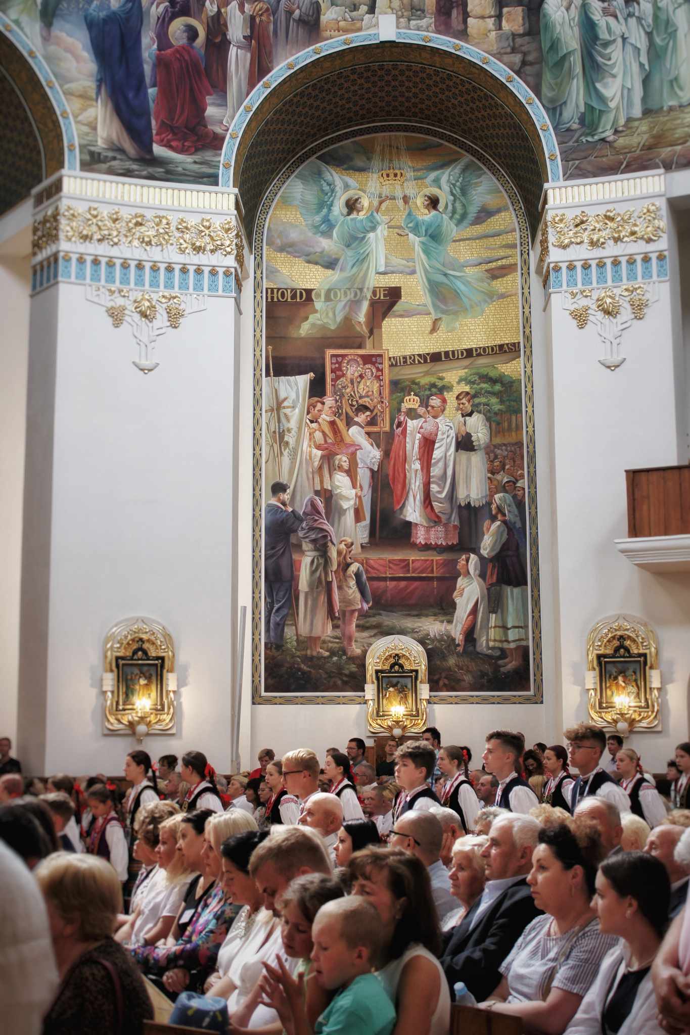 Wnętrze kościoła z widocznym obrazem na ścianie przedstawiającym scenę religijną. W sali zgromadzeni, głównie starsi mężczyźni i kobiety, ubrani w formalne stroje.