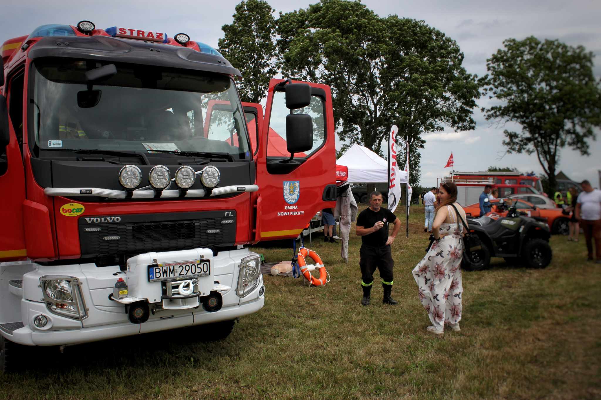 Na zdjęciu przedstawiono czerwony samochód strażacki marki Volvo i drugi pojazd, z udziałem osób stojących w pobliżu. Na trawniku leży sprzęt ratowniczy.