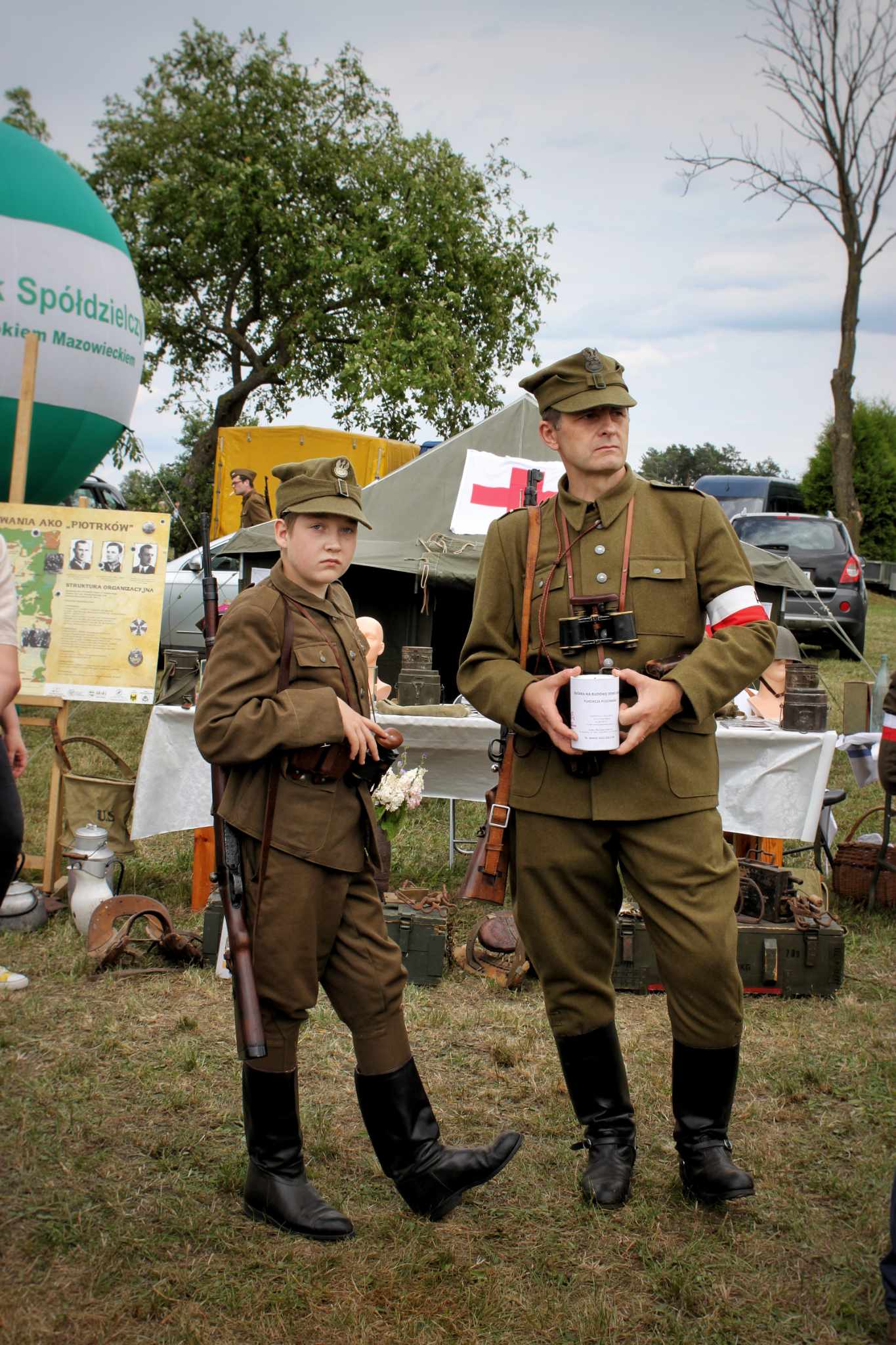 Na zdjęciu przedstawiono dwoje osób w mundurach wojskowych, młodsza trzyma broń, a starsza aparat fotograficzny. W tle znajduje się namiot z symbolem krzyża i biało-czerwonym paskiem oraz inne elementy krajobrazu.