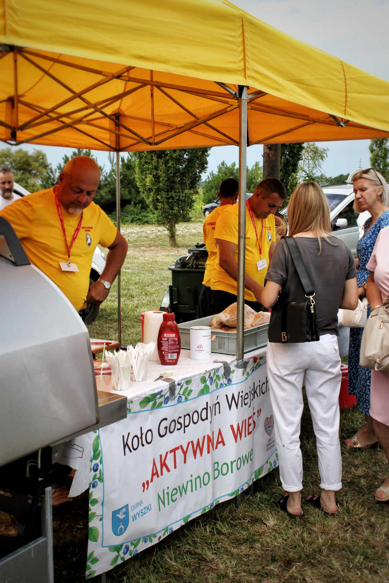 Na zdjęciu przedstawiono ladę z przekąskami i napojami obsługiwaną przez wolontariuszy w żółtych koszulach. W tle znajduje się trawnik i grupa osób czekająca w kolejce.