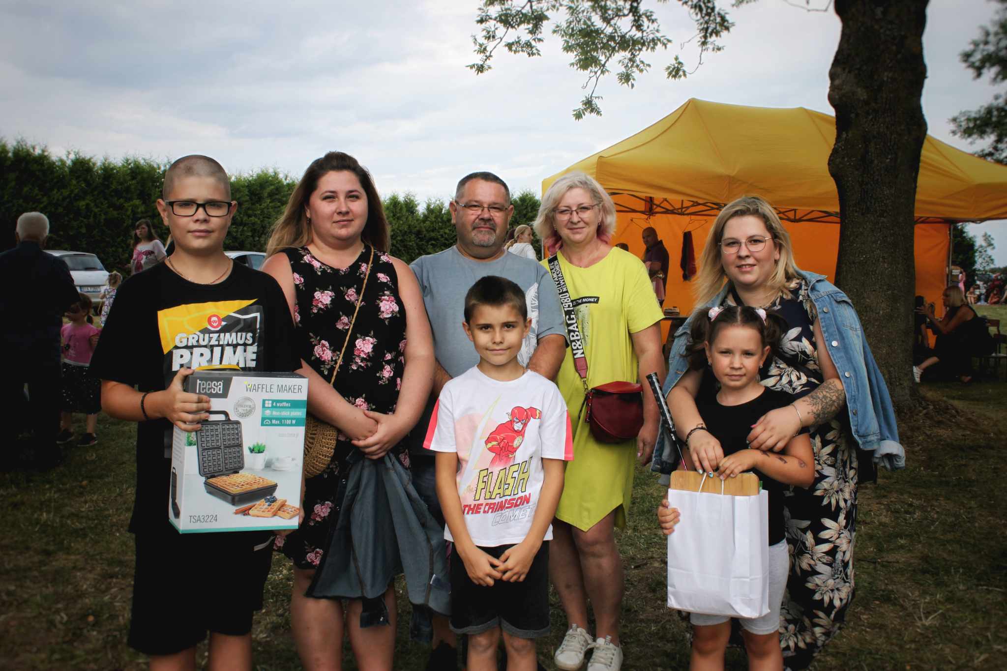Zdjęcie przedstawia rodzinę podczas wydarzenia. Osoby są ubrane w odzież letnią i trzymają różne przedmioty, takie jak pudełko z produktem i torby papierowe.