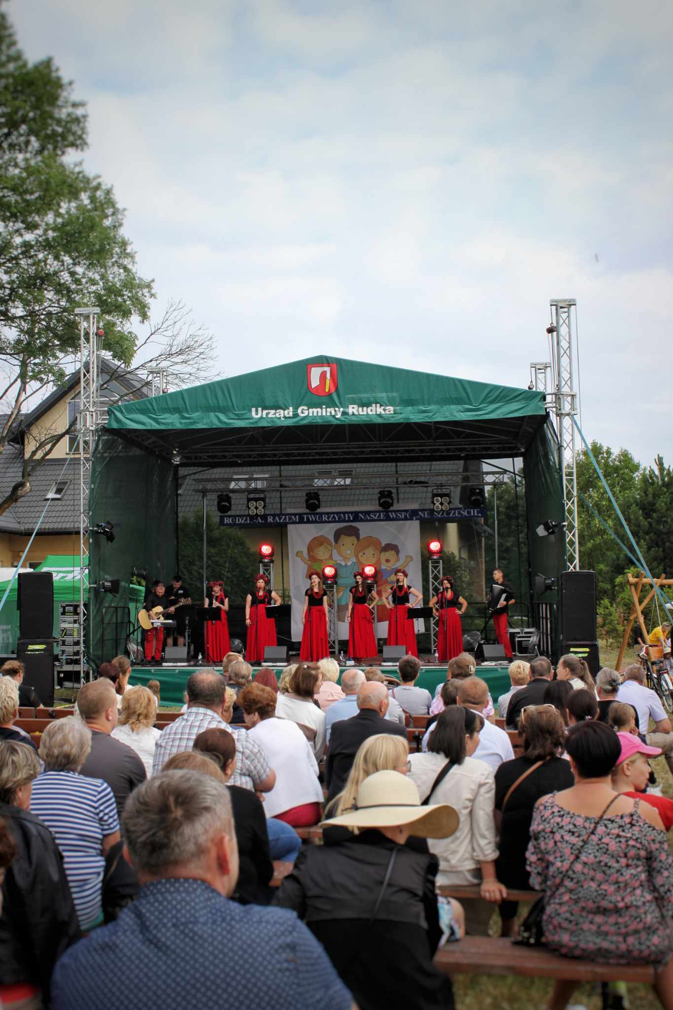 Na scenie występuje grupa muzyków w czerwonych strojach, otoczona przez publiczność. W tle widoczny namiot sceniczny z logo. Wydarzenie odbywa się na świeżym powietrzu.