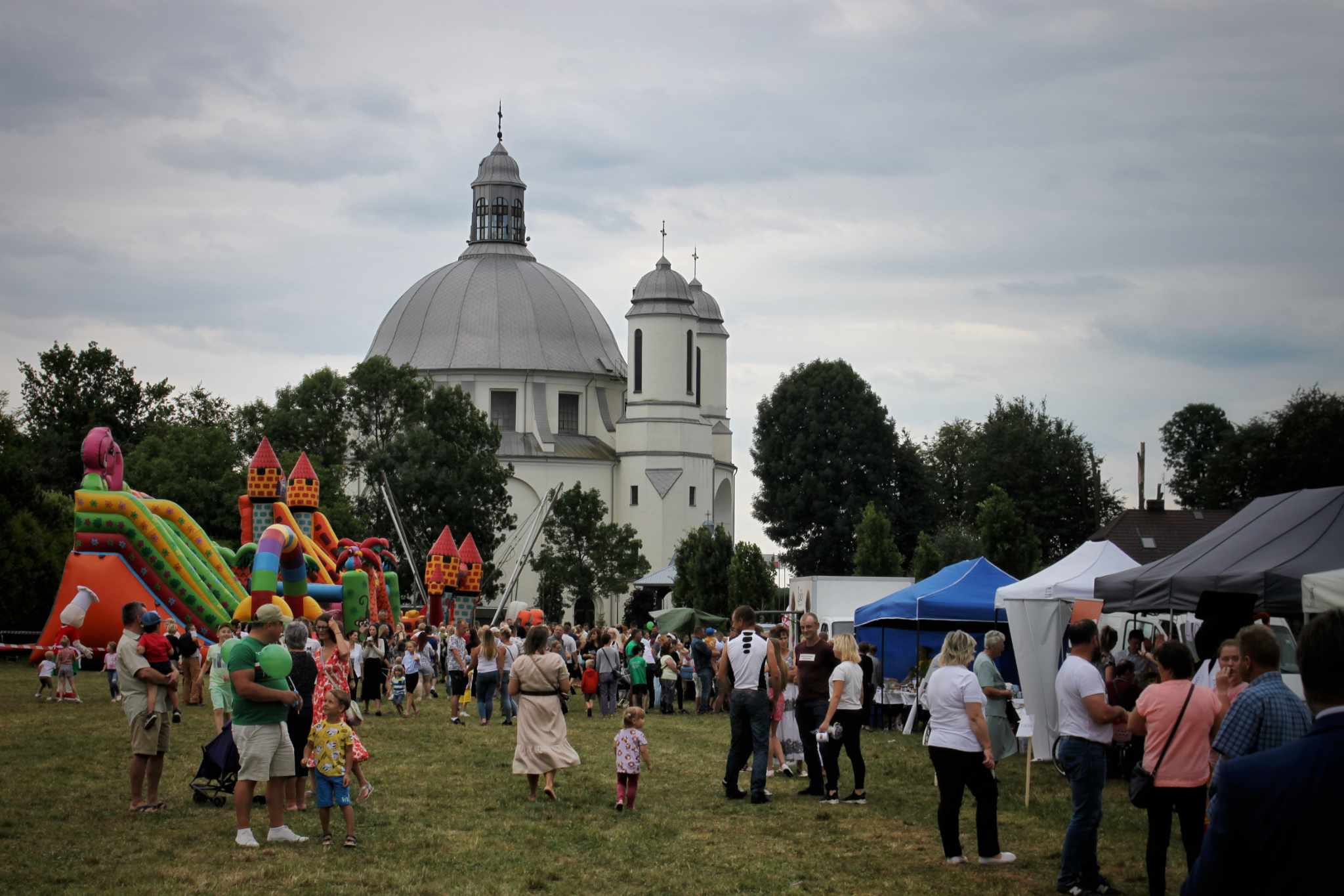Na trawniku zgromadzeni ludzie, przed kościołem z kopułą i dmuchanymi atrakcjami dla dzieci. Widać również stoiska.