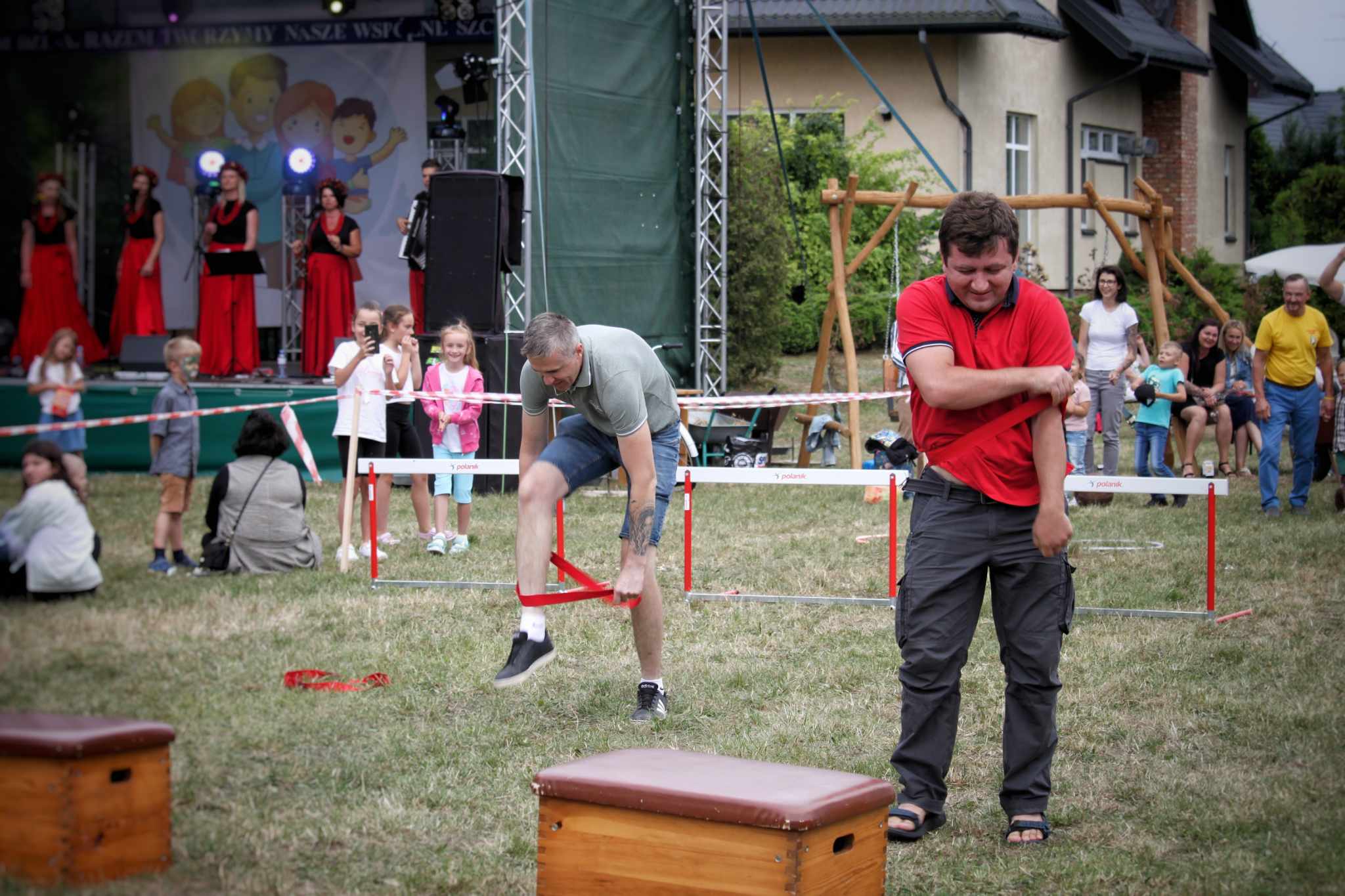 Dorośli uczestnicy konkurencji sportowej na trawniku podczas V Dnia Rodziny; w tle scena i publiczność.
