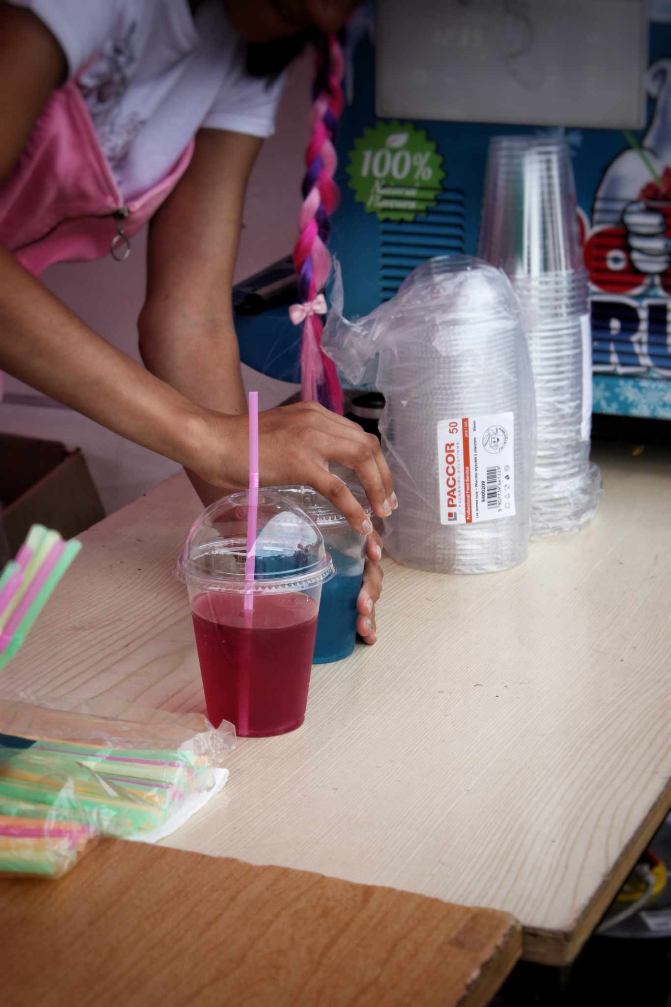 Na stole znajduje się szklanka z różowym napojem i słomką, obok kubki plastikowe oraz pojemnik na słomki. W tle fragment plakatu z drinkiem.
