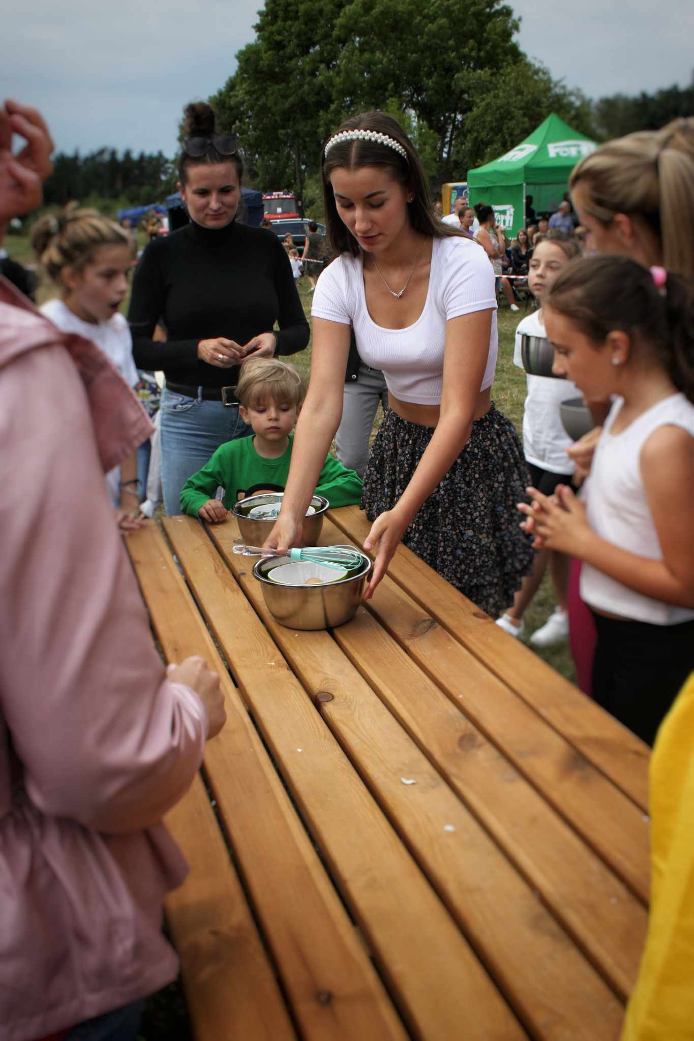 Na stole znajdują się naczynia z płynem, a wokół nich zgromadzeni ludzie. W tle zielony namiot i drzewa.